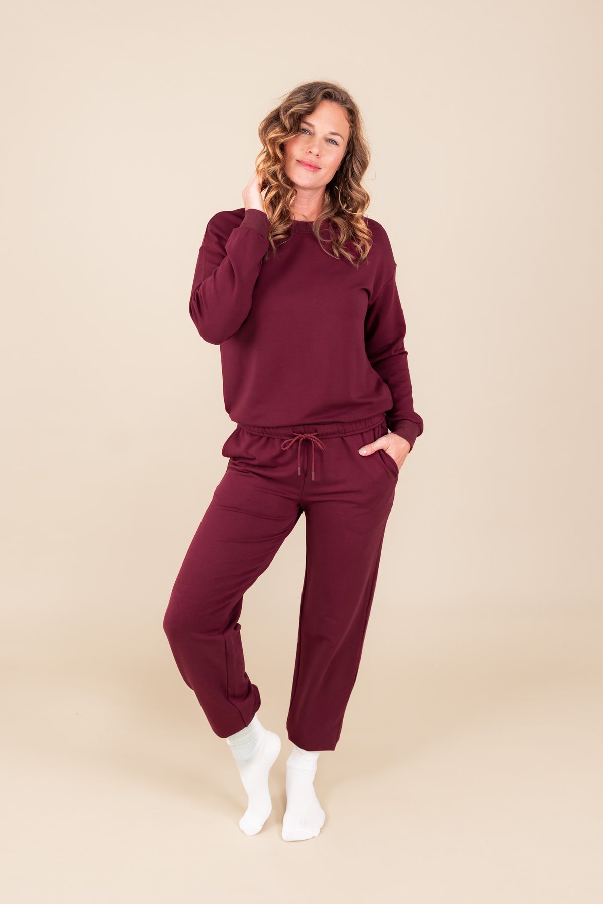 Modal posing in Women's Jogger Pant in Burgundy color under similar color sweatshirt and white socks on a beige background