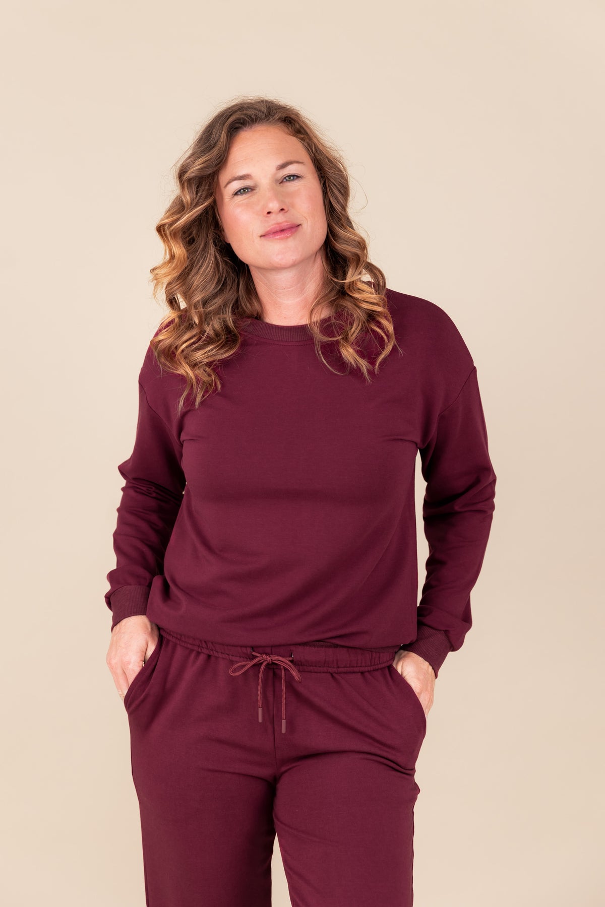 Woman wearing Women's Sweatshirt in Burgundy color over women's jogger pants in burgundy on a beige background