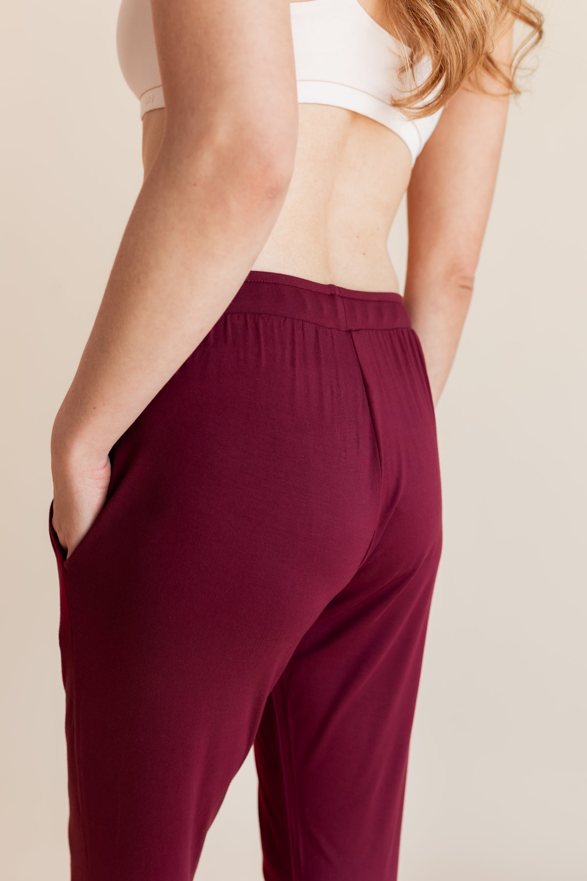 Model showcasing back side of Women's Jogger Pants in Burgundy under white bralette on a beige background