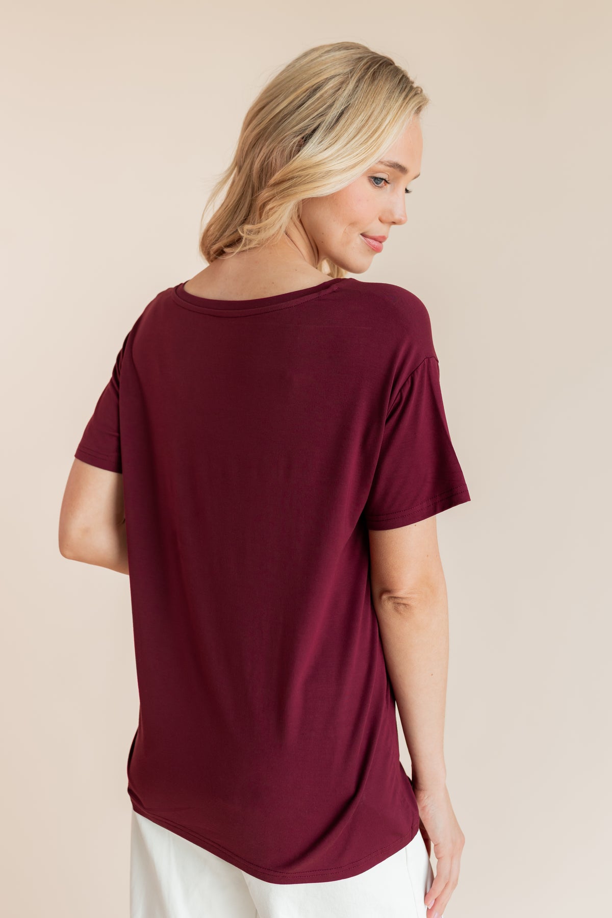Model showcasing backside of Women's Oversized V-Neck Tee in Burgundy over white pants on a beige background