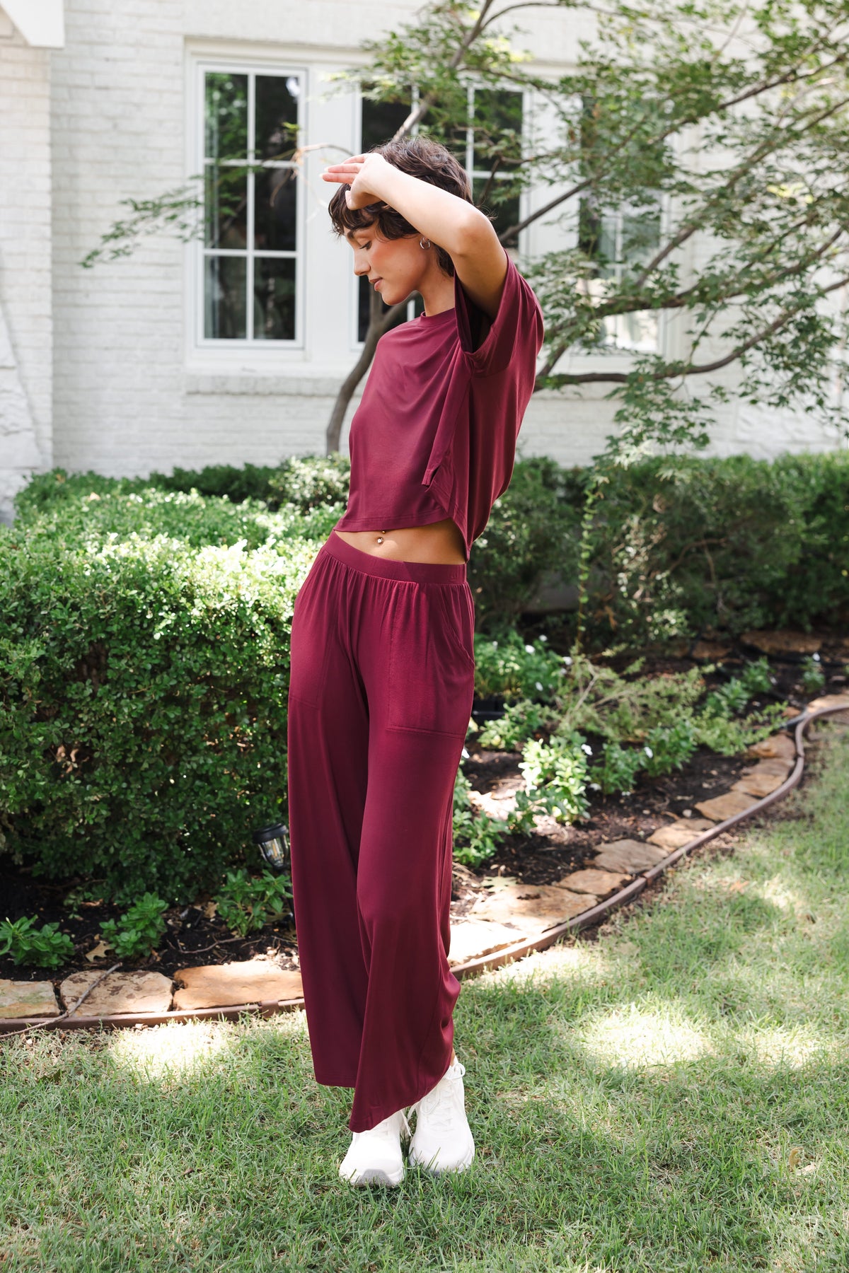 Female model standing in garden wearing the Women's Wide Leg Pant in Burgundy and matching short sleeve tee over white shoes