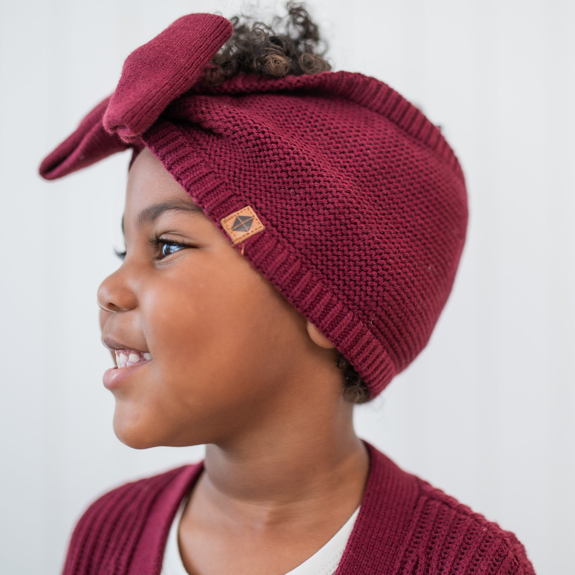 Side view of the Chunky Knit Bow Headband in Burgundy with the Kyte Baby Vegan leather tag showing