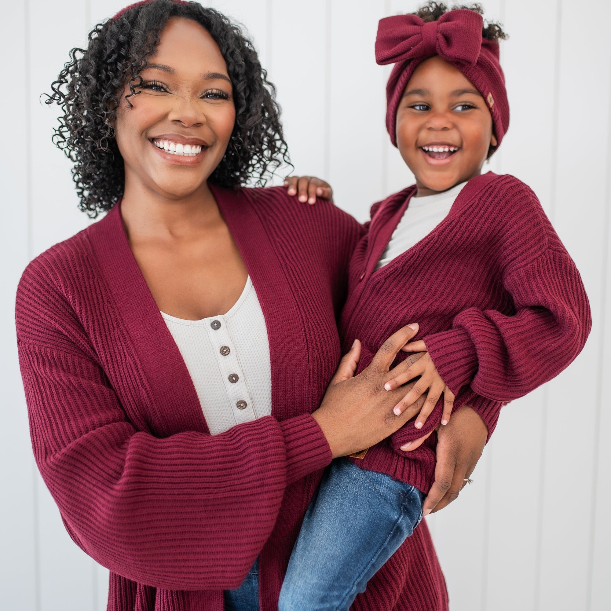 Mother holding daughter both matching in Burgundy Chunky knit sweaters. Young girl has the Chunky Knit Bow Headband in Burgundy paired with her outfit