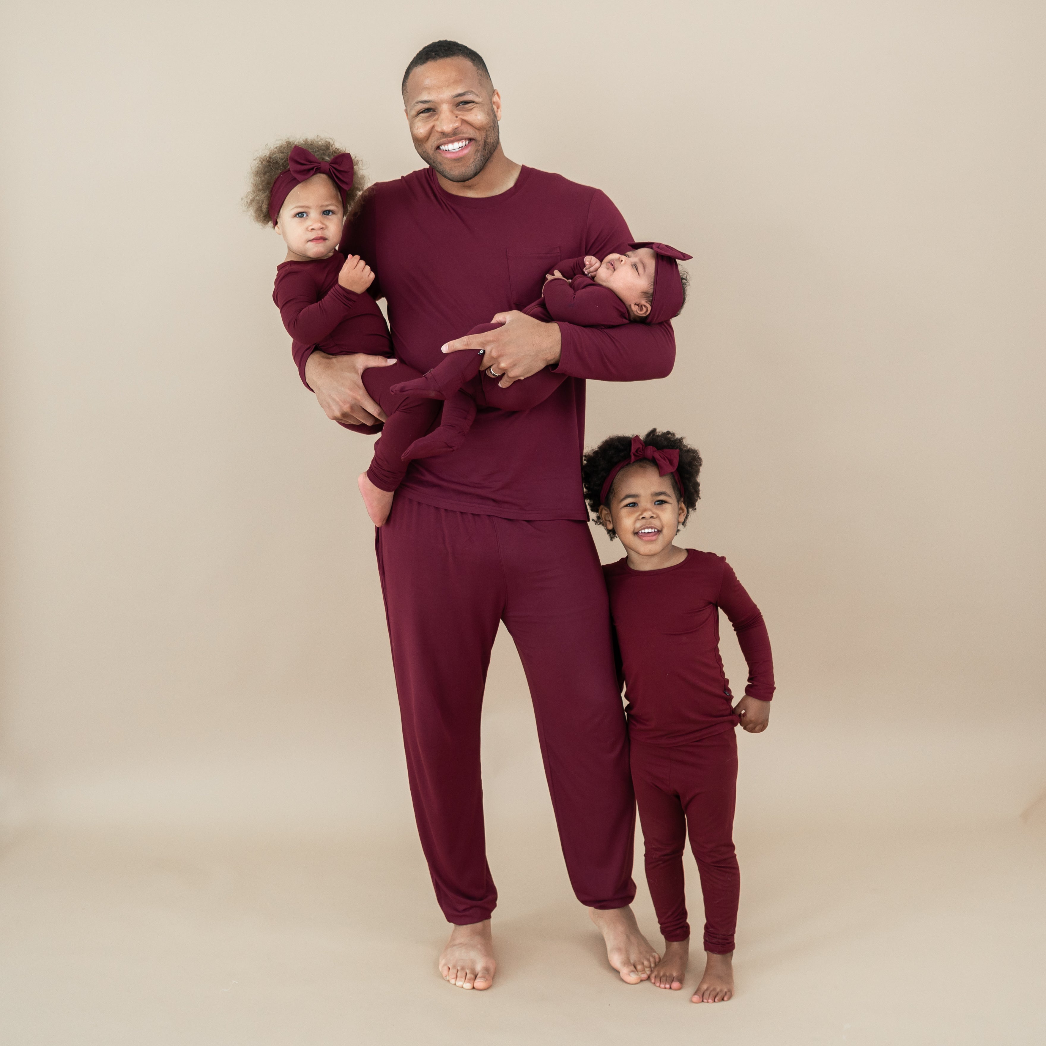 Family matching in Burgundy. Father holding younger two daughters with his oldest daughter standing right next to him. Pictured are the men's joggers, zippered footie and toddler pajama sets