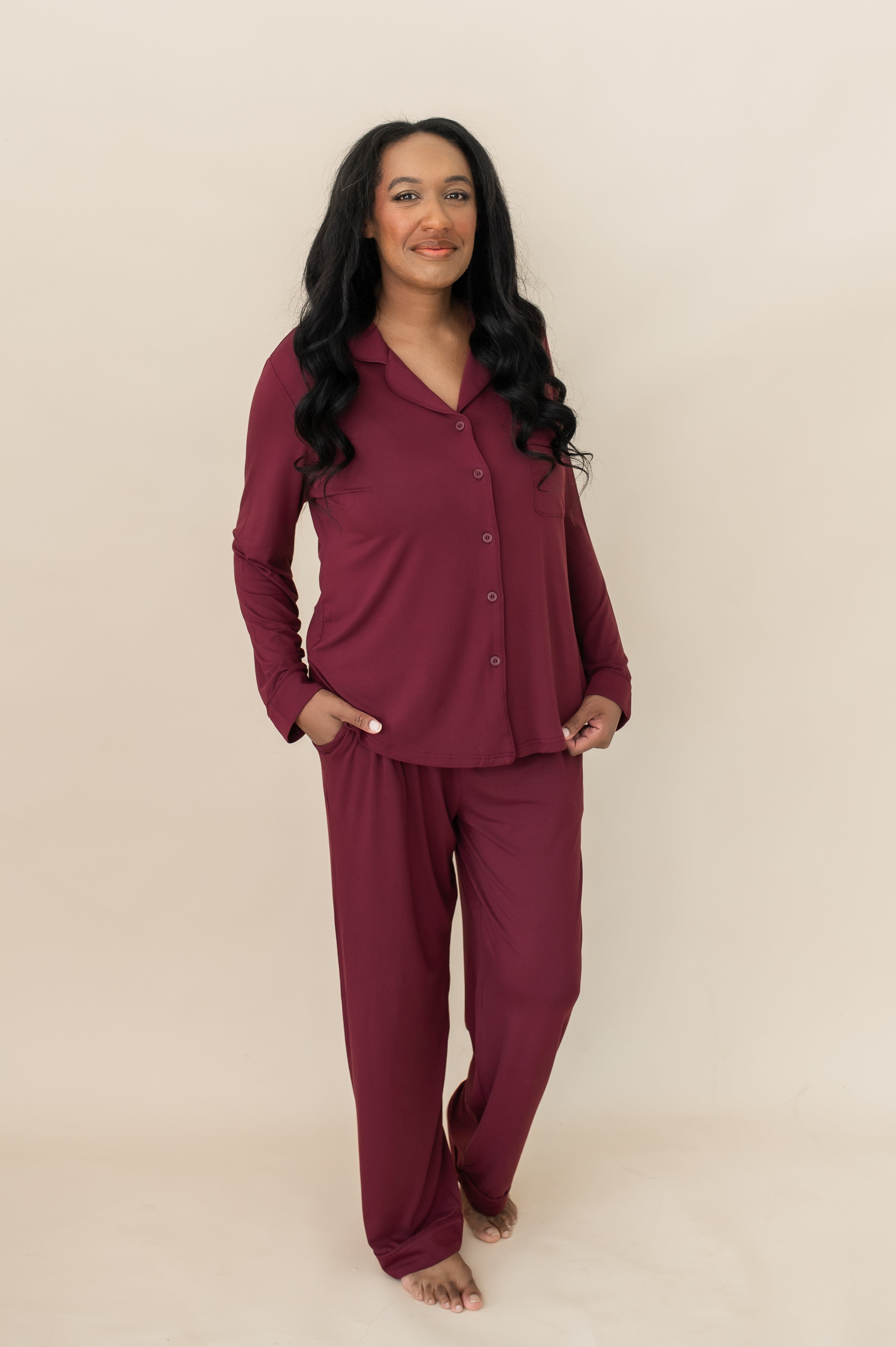 Female wearing the Long-Sleeved Women's Pajama Set in Burgundy with Burgundy Trim with one hand in the pants pocket and the other holding the bottom hem of the shirt standing in front of a light neutral background
