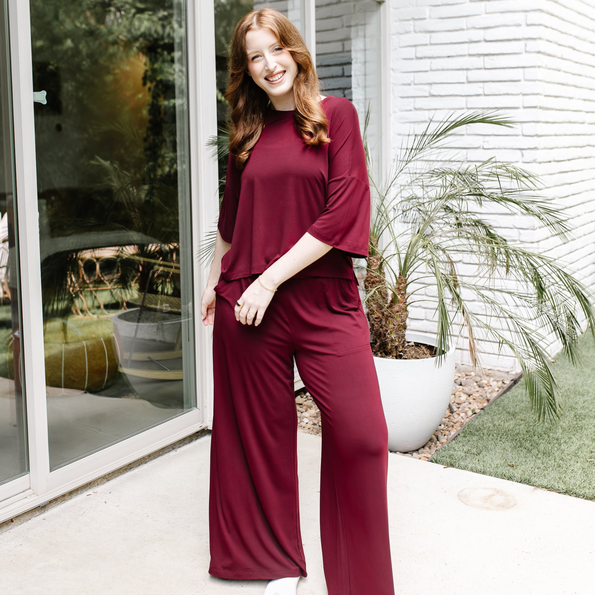 Female model standing in front of a glass window on a white brick house wearing the Women's Wide Leg Pant in Burgundy and matching short sleeve tee