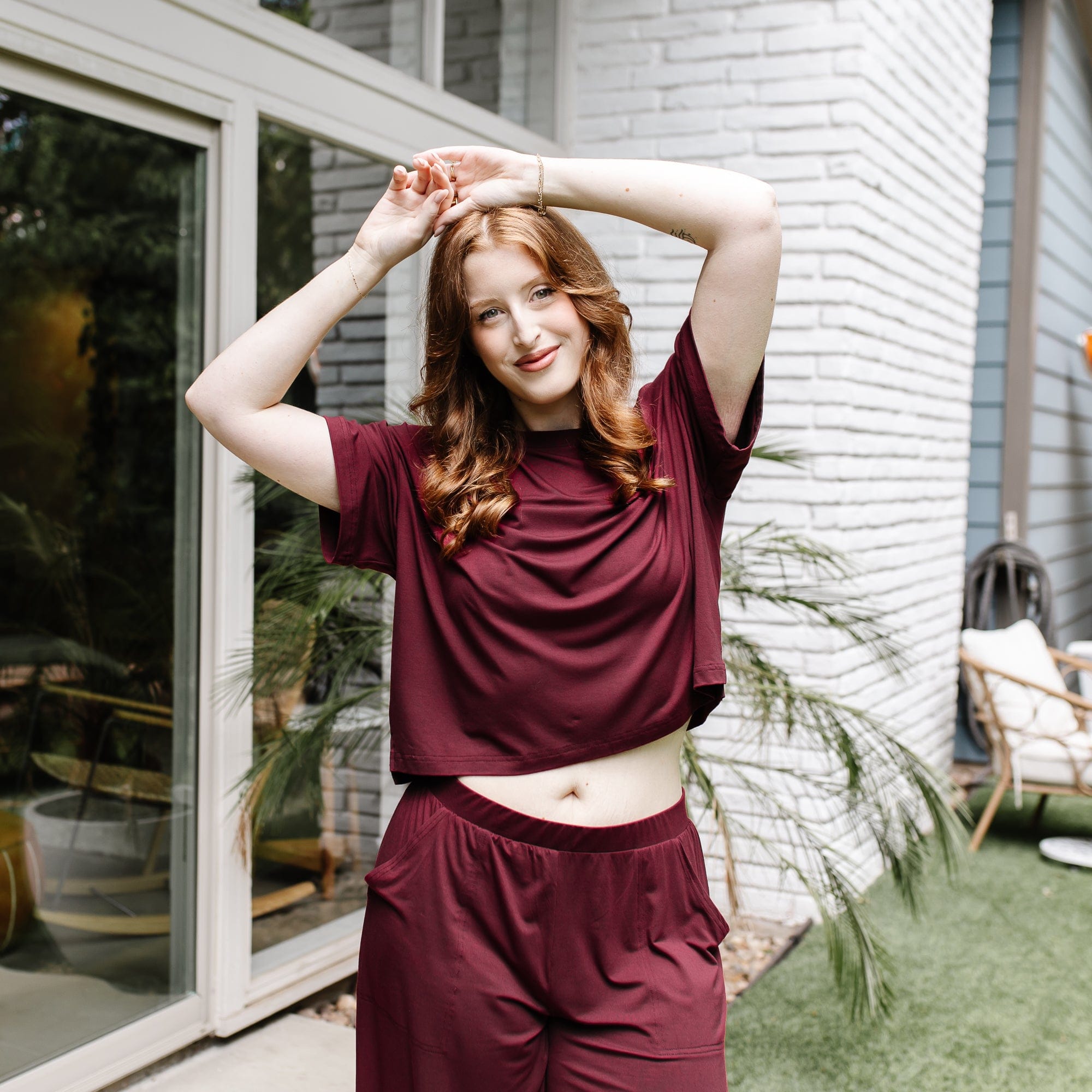 Female model standing with her hands stacked on her head wearing the Women's Short Sleeve Tee in Burgundy in front of a white brick house