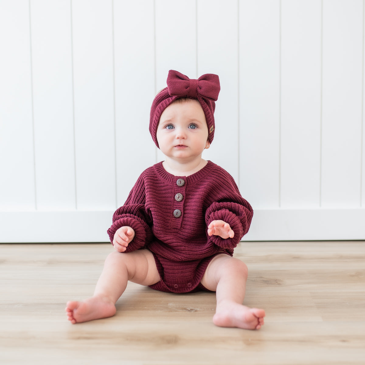 Toddler sitting wearing the Chunky Knit Romper in Burgundy with matching chunky knit bow headband