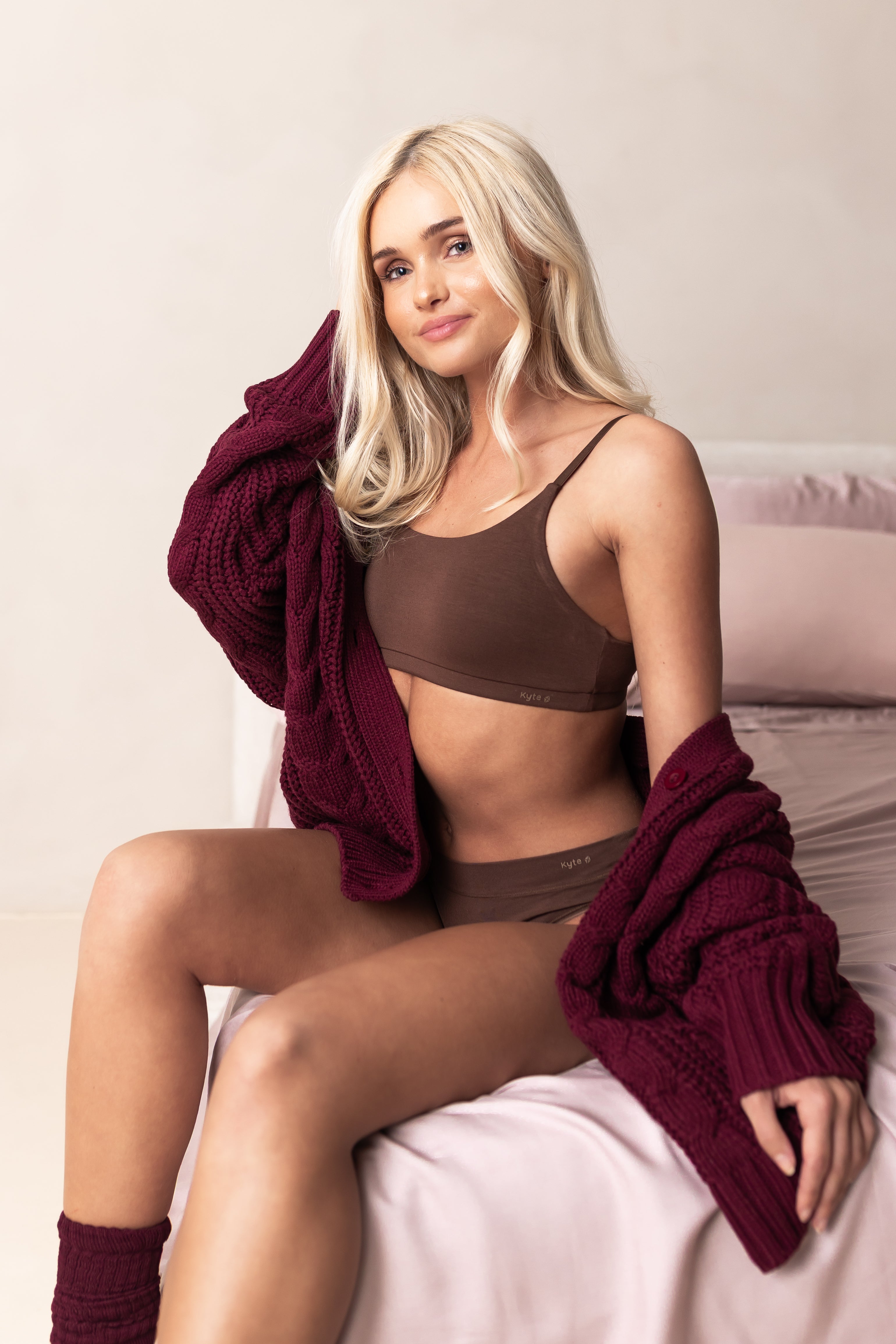 Woman in a brown scoop neck bralette and underwear set with a maroon cardigan on a light background