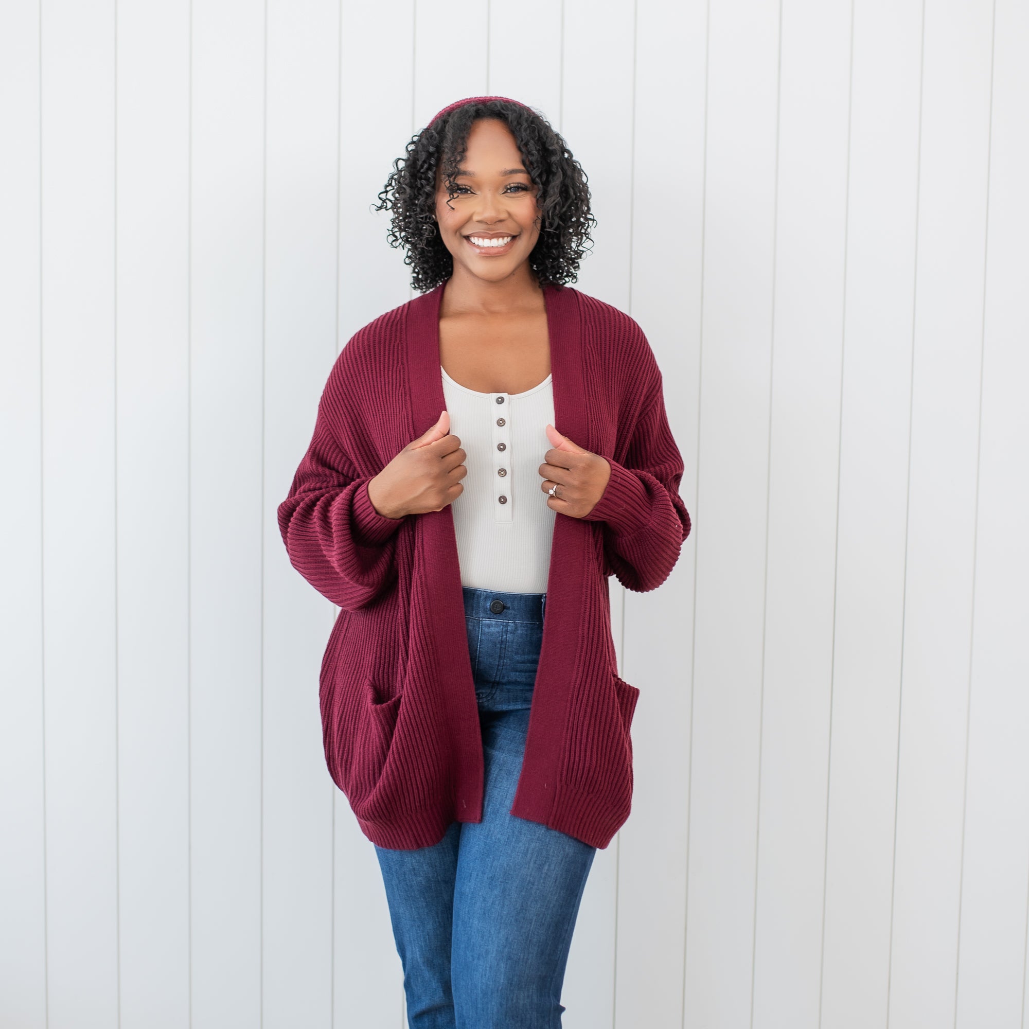 Female standing in front of a white paneled wall wearing the Chunky Knit Women's Oversized Cardigan in Burgundy and medium wash jeans