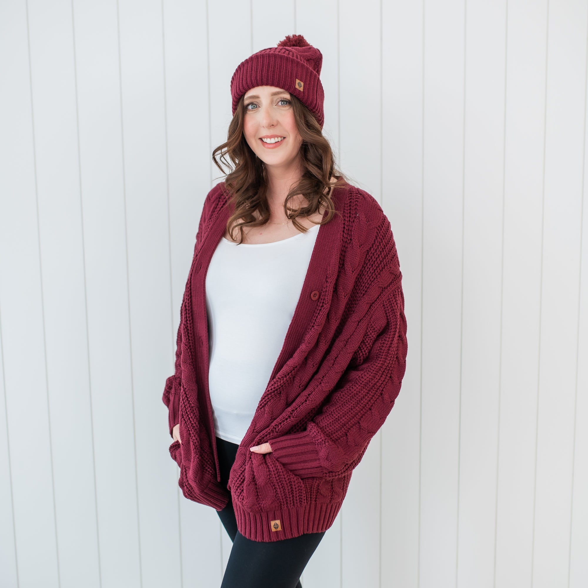 Smiling female model standing with hands in the pocket of the Cable Knit Adult Cardigan in Burgundy paired with a matching chunky knit beanie