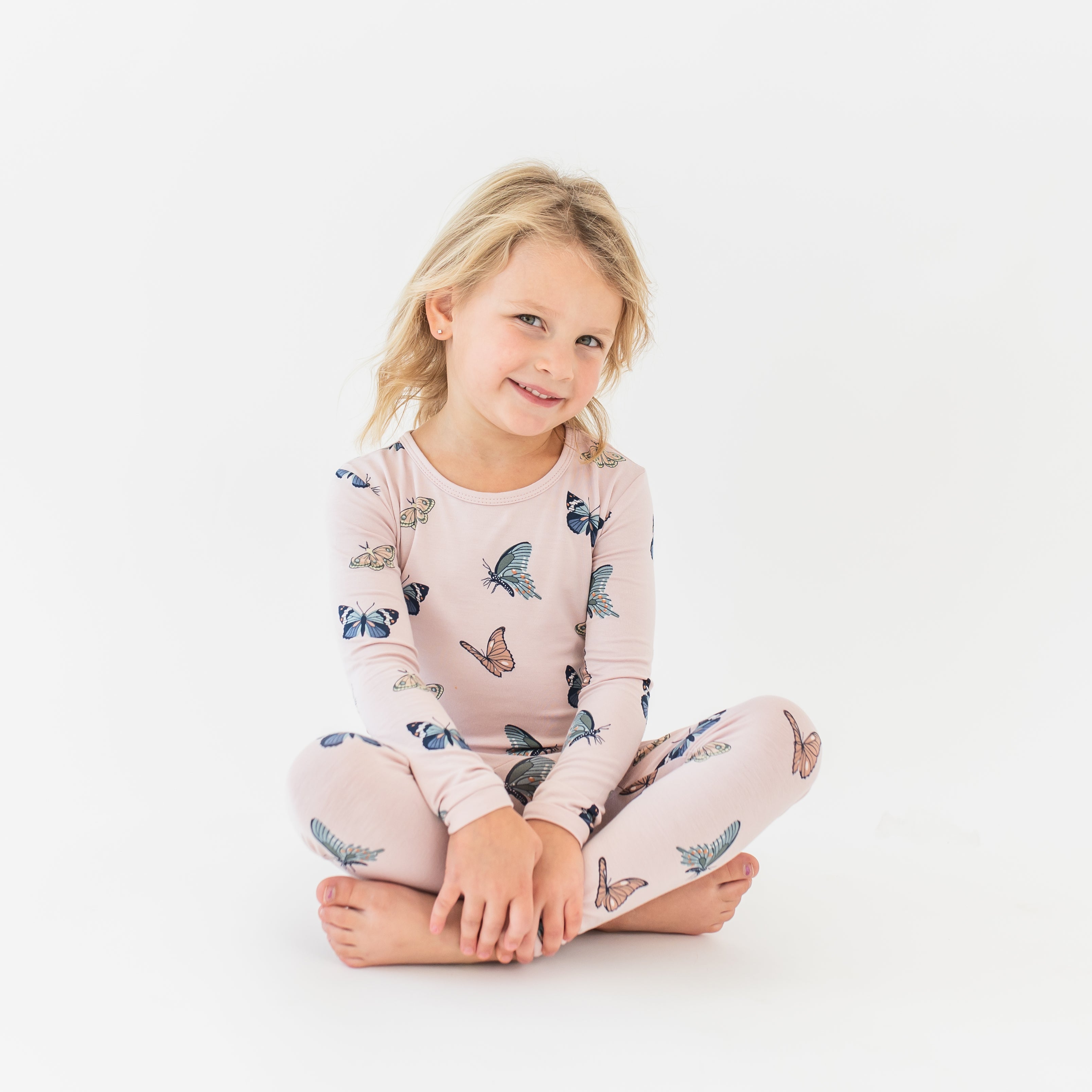 Young girl sitting on the floor cross legged wearing the Long Sleeve Pajamas in Blush Butterfly