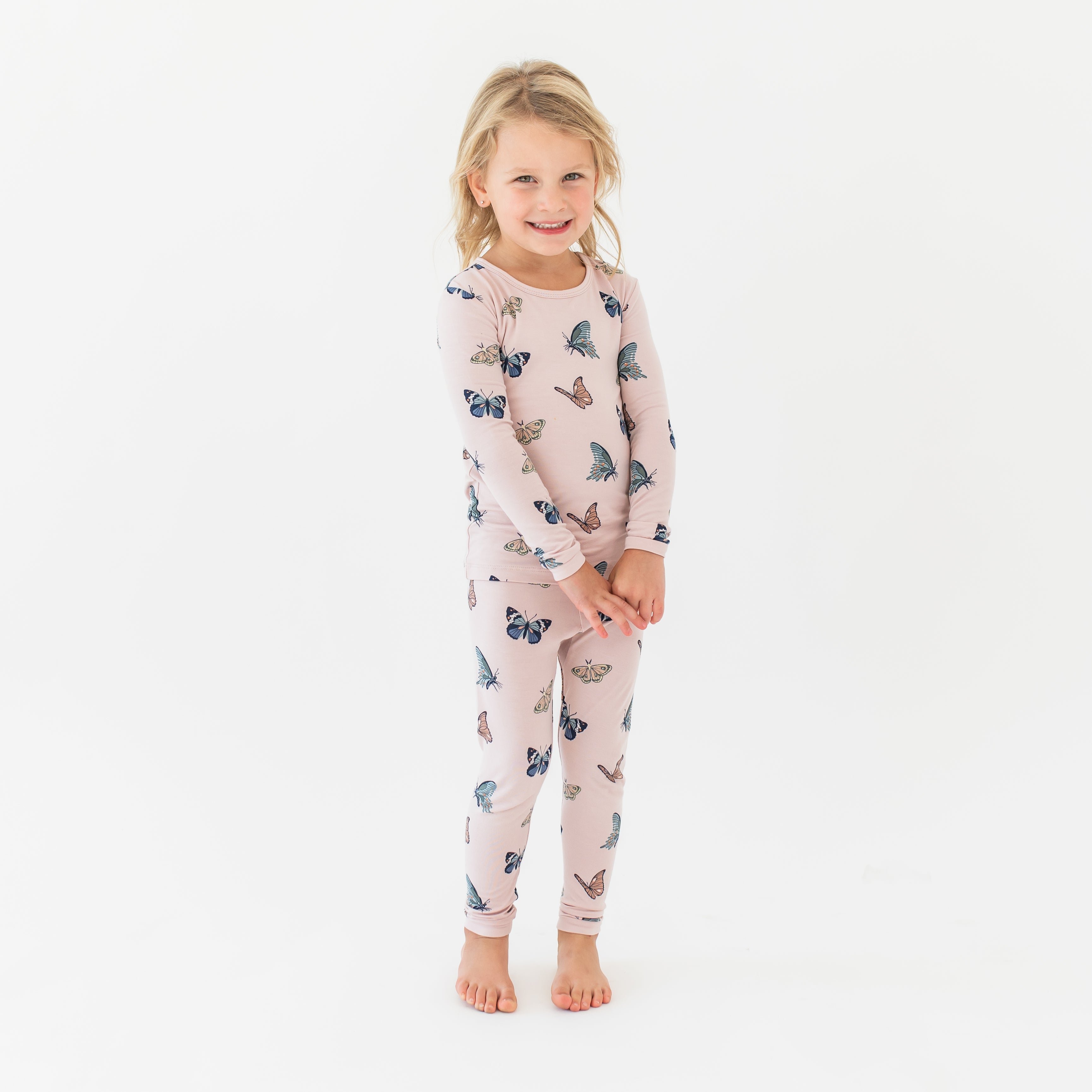 Young smiling girl standing with her hands clasped together wearing the Long Sleeve Pajamas in Blush Butterfly