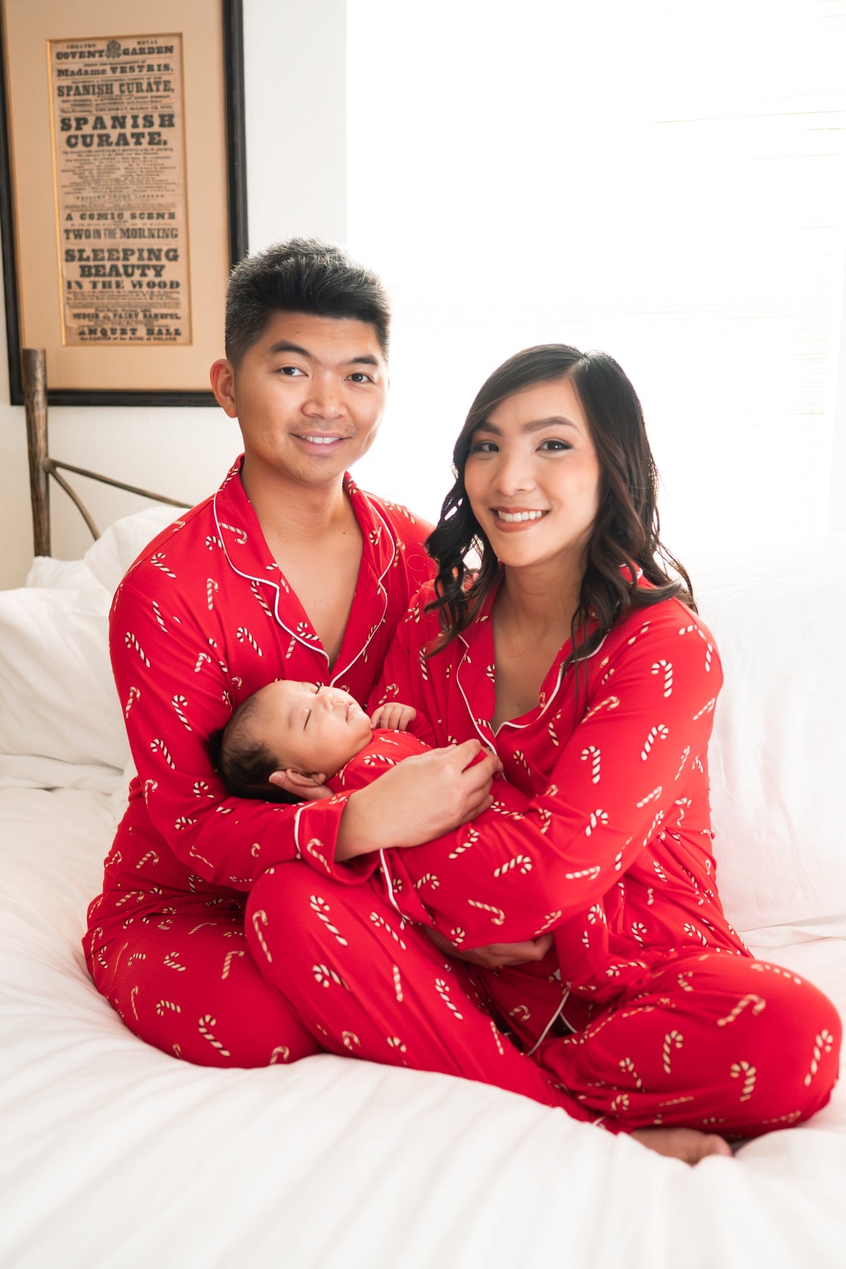 Family of three sitting on a bed matching in Candy Cane. Pictured are the Women's and Men's Long Sleeve Pajama Set in Candy Cane and zippered footie