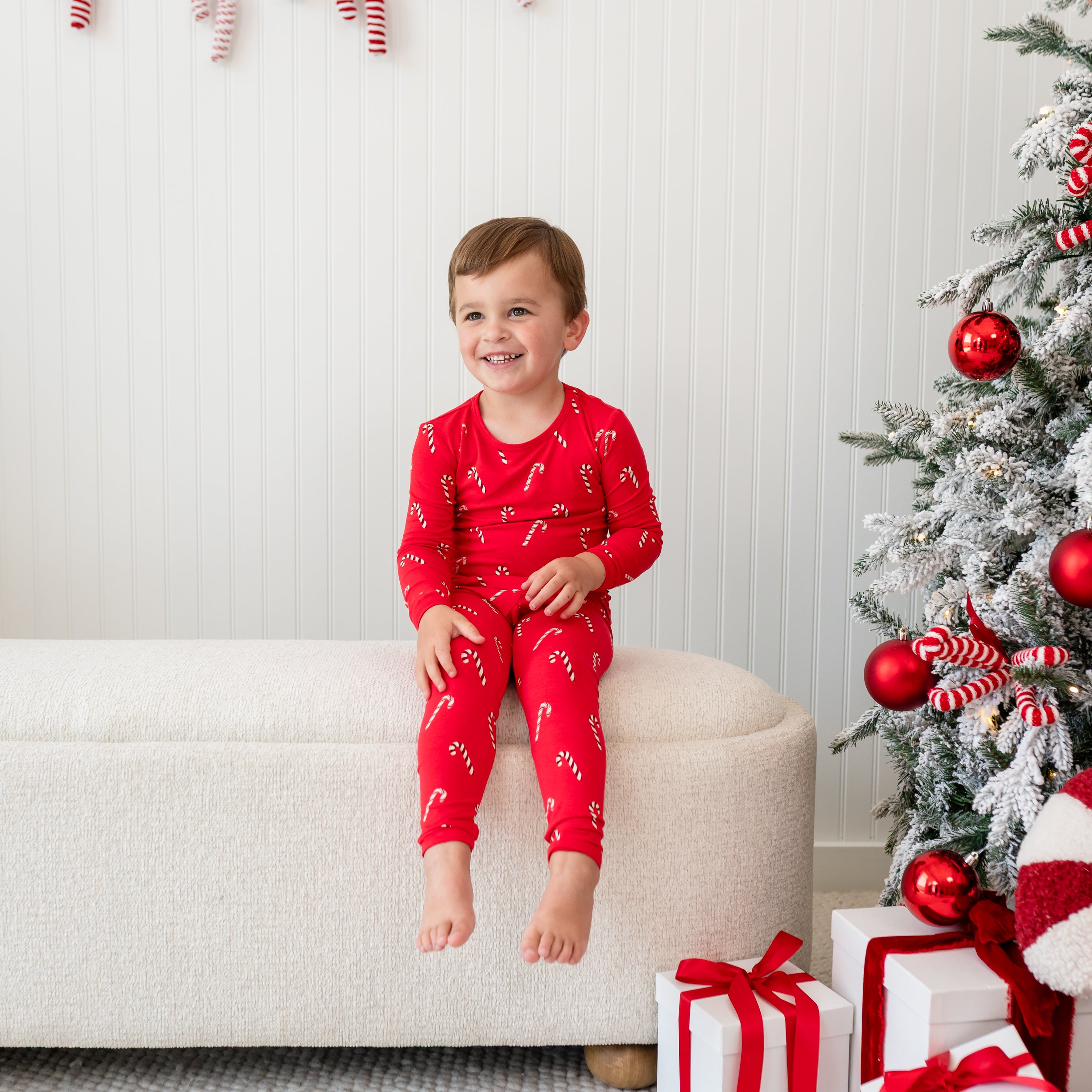 Young boy sitting on a cream ottoman beside a decorated Christmas tree wearing the Long Sleeve Pajamas in Candy Cane