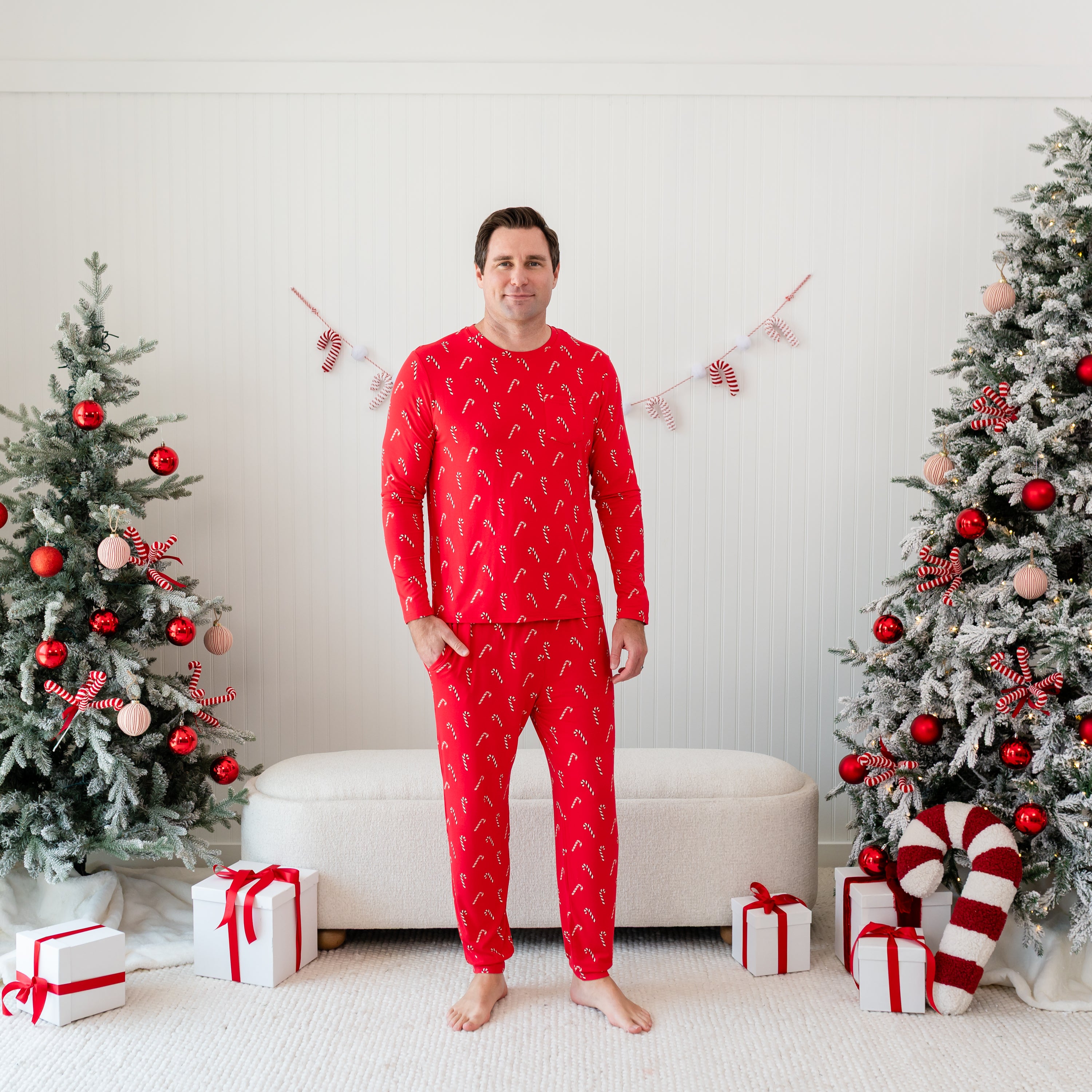 Male model standing in front of a cream ottoman between two decorated Christmas trees wearing the Men's Jogger Set in Candy Cane with one hand in the jogger pants pocket