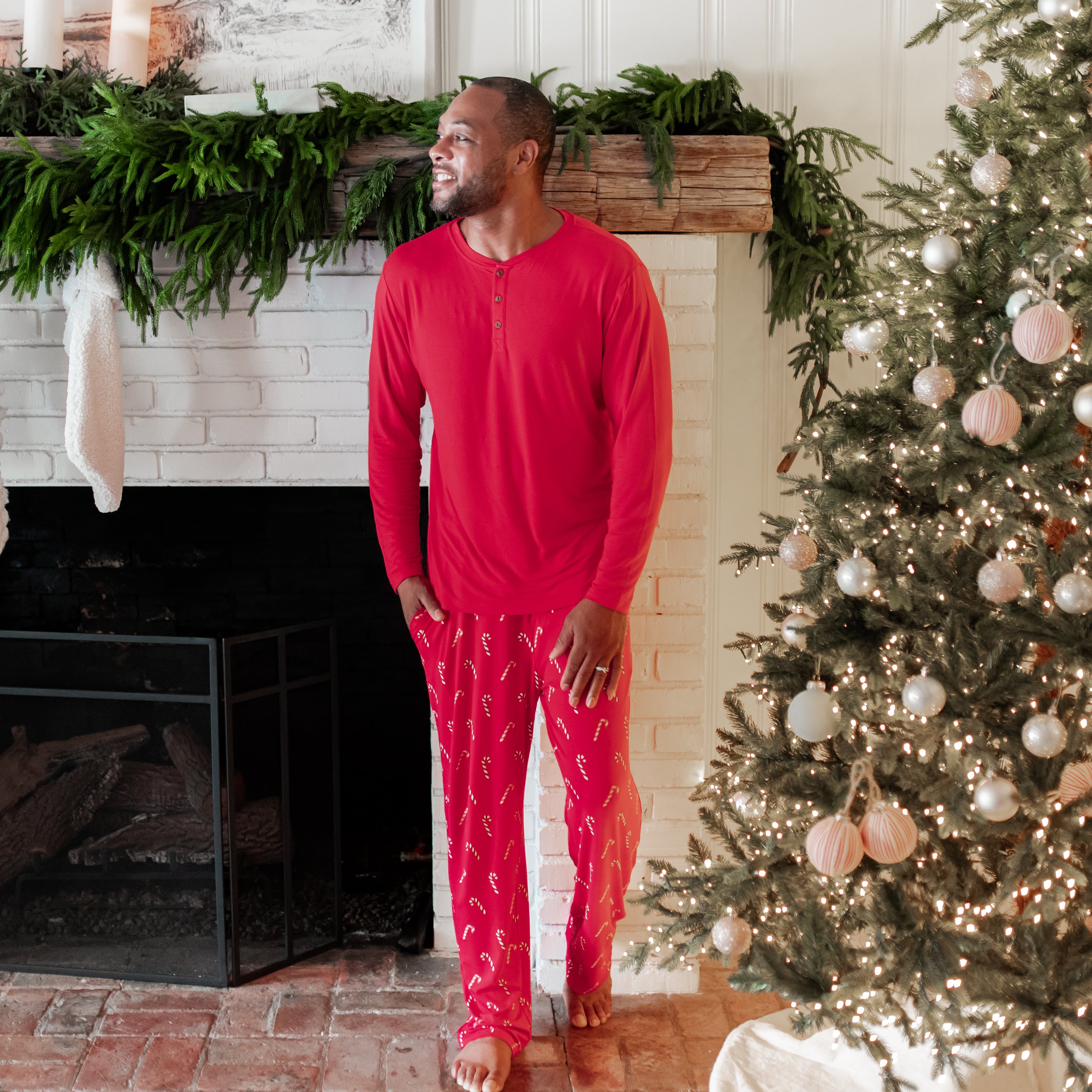 Male model walking looking off to the side in front of a white brick fireplace wearing the Men's Lounge Pants in Candy Cane and Cardinal men's Henley top