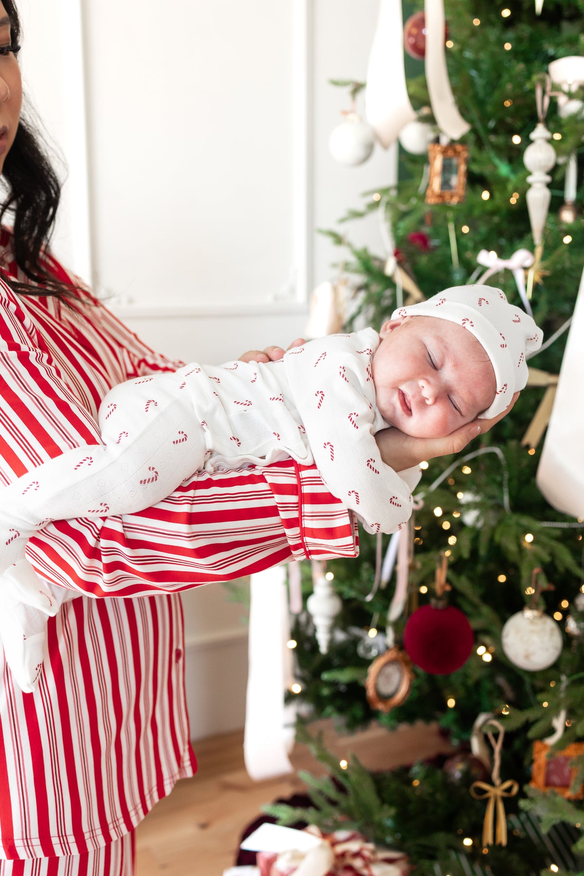 Mommy wearing striped red and white long sleeve pajama set holding her sleeping baby wearing Pointelle Take Me Home Set with Cap in Candy Cane print near a Christmas tree