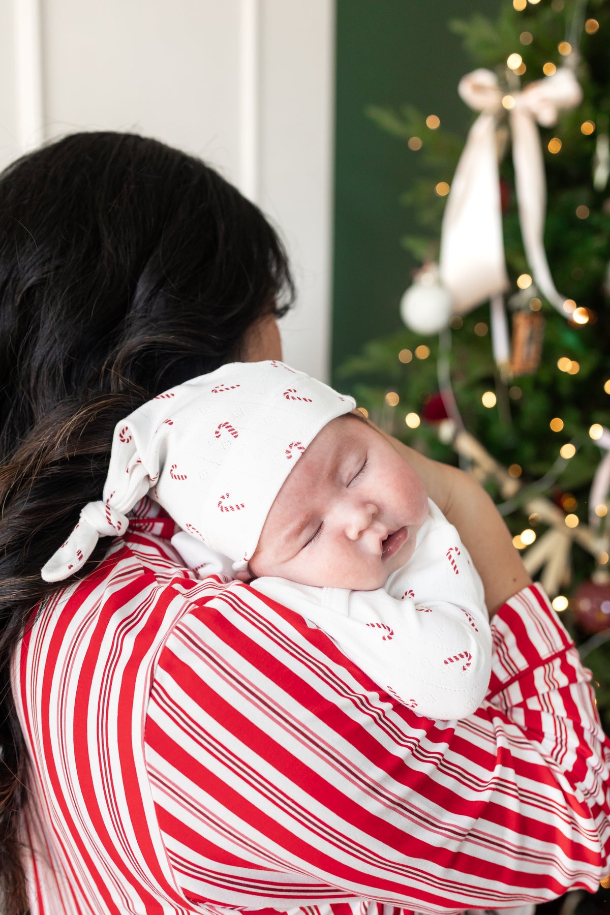 Mommy wearing striped red and white long sleeve pajama set holding her sleeping baby on her shoulder wearing Pointelle Take Me Home Set with Cap in Candy Cane print near a Christmas tree