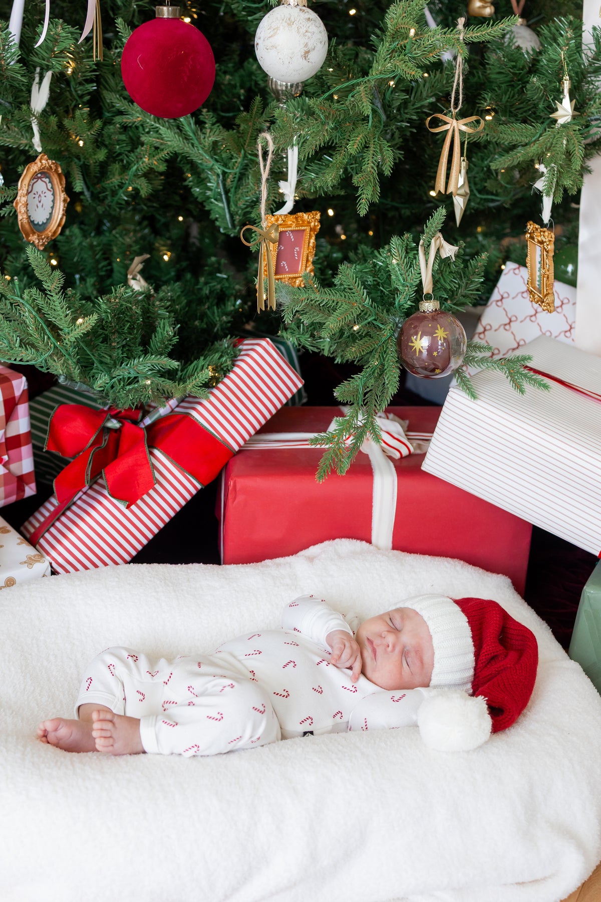Baby sleeping wearing Pointelle Kimono Set in Candy Cane and red hat on a white sheet near a Christmas tree and presents