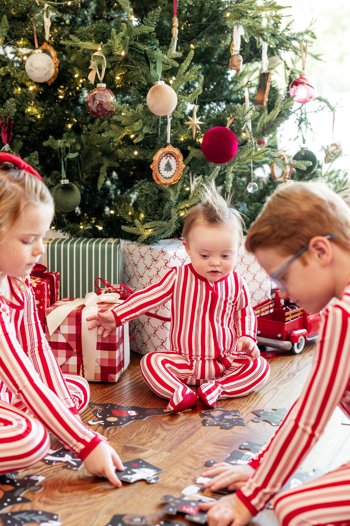 Three children wearing matching red and white striped Christmas pajamas sit on the floor in front of a decorated Christmas tree, with wrapped gifts and a red toy truck nearby.