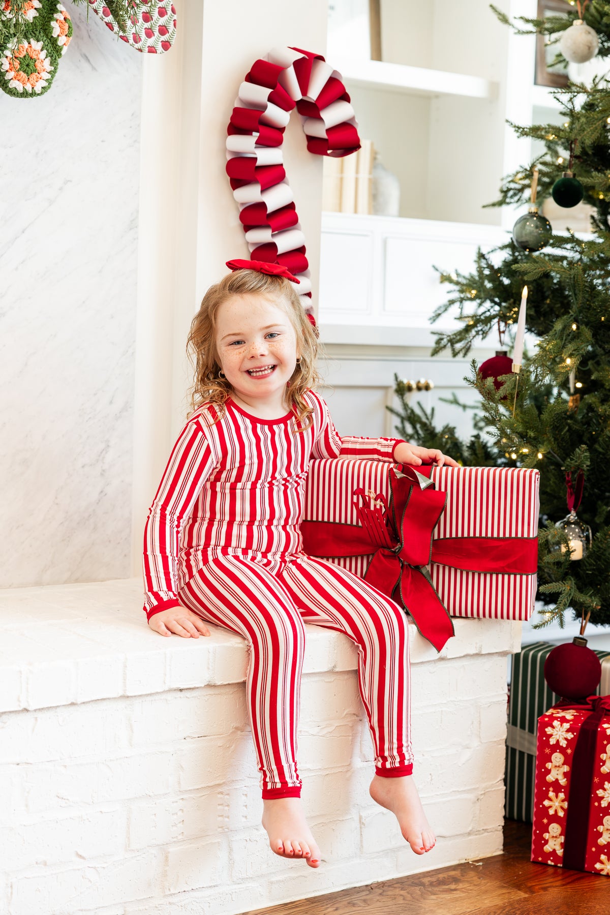 A young girl with blonde hair sits smiling on a white brick fireplace mantel, wearing a red and white vertically striped long-sleeve pajama set. She is holding a large red and white striped gift box with a red bow.