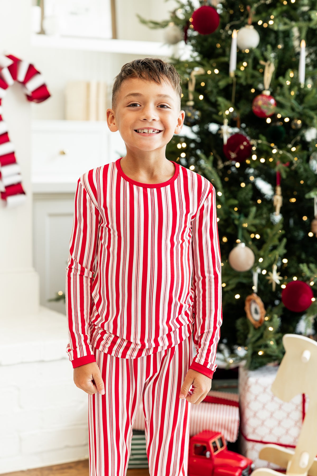 Boy smiling in Long Sleeve Pajamas in Peppermint Stripes print with Christmas tree in the background