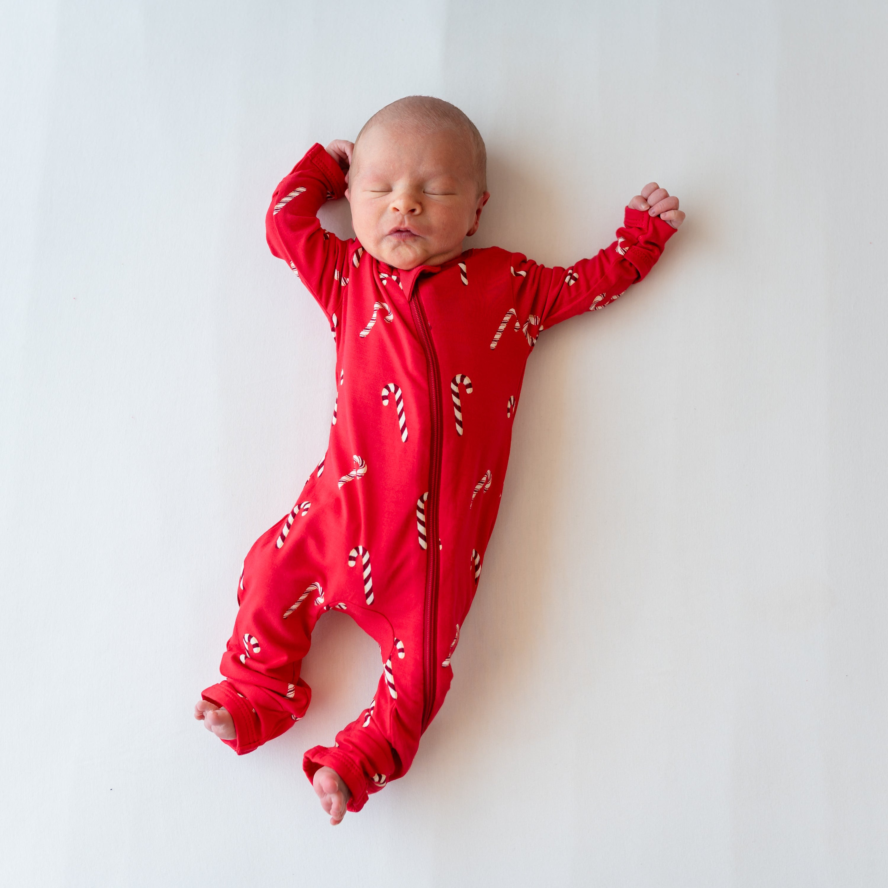Sleeping newborn laying down wearing the Zippered Romper in Candy Cane on a white blanket