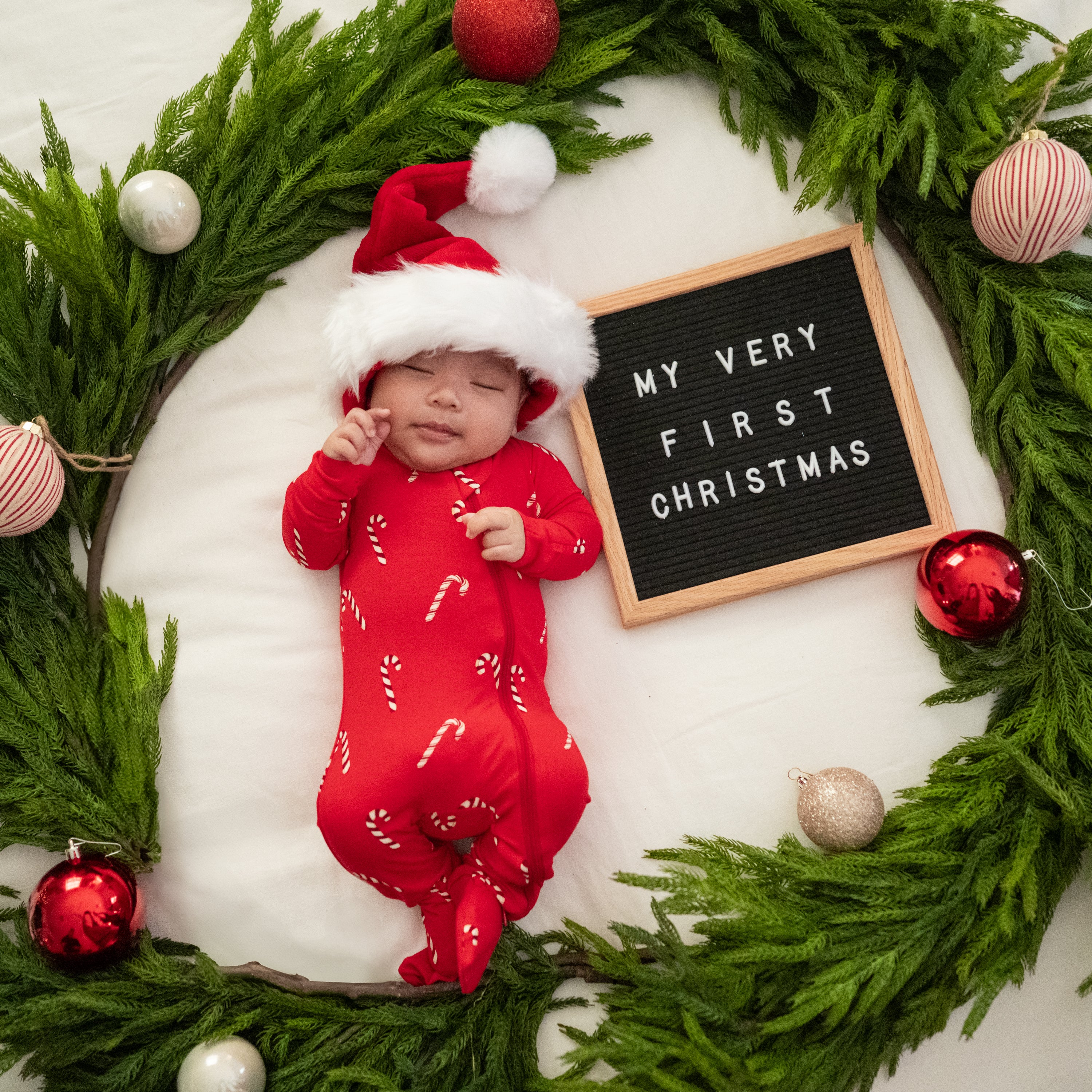Sleeping infant wearing the Zippered Footie in Candy Cane laying on a blanket with a Christmas wreath around hem and a word board saying 'My very first Christmas'