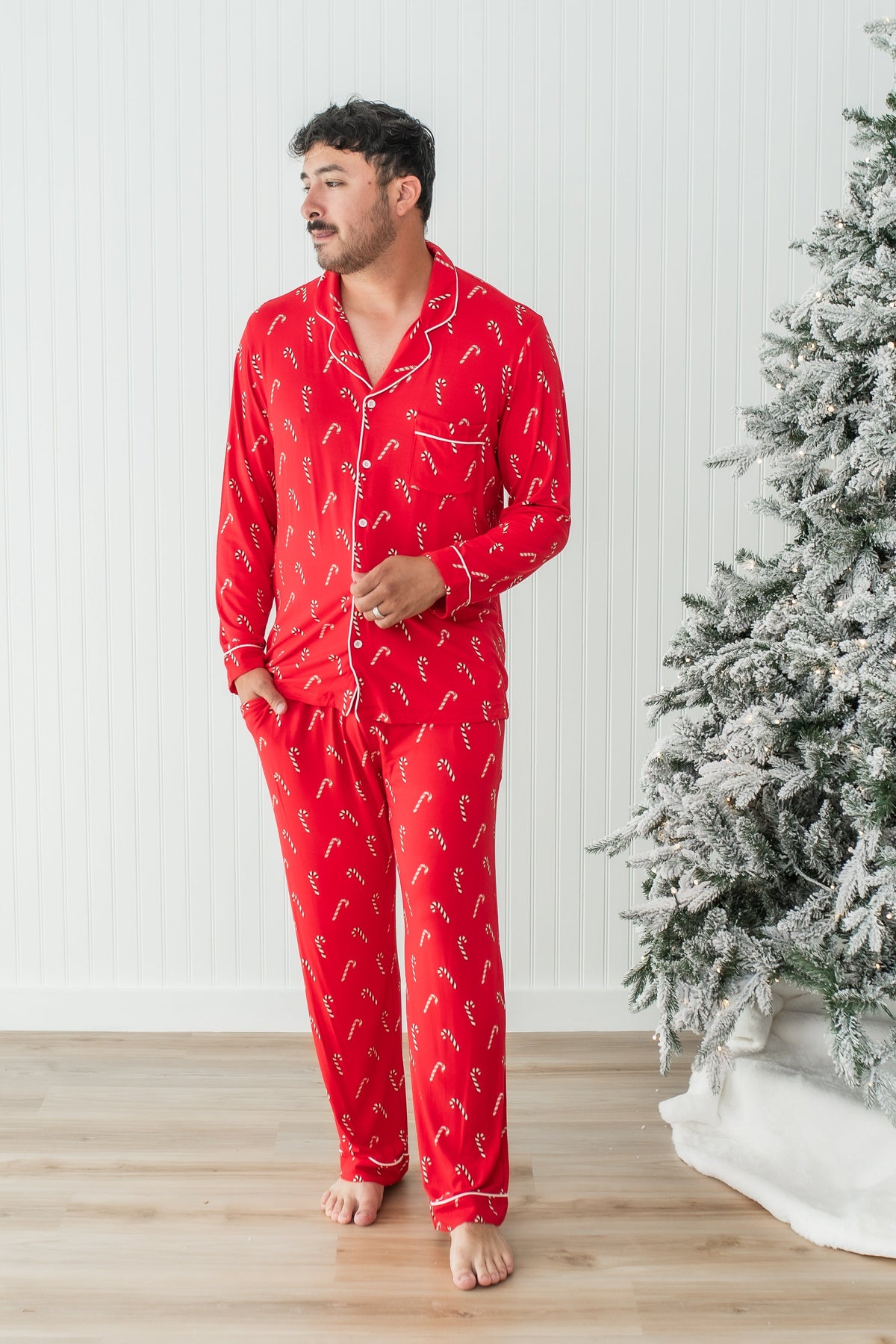 Male model wearing the Men's Long Sleeve Pajama Set in Candy Cane standing in front of a white paneled wall beside a frosted tree