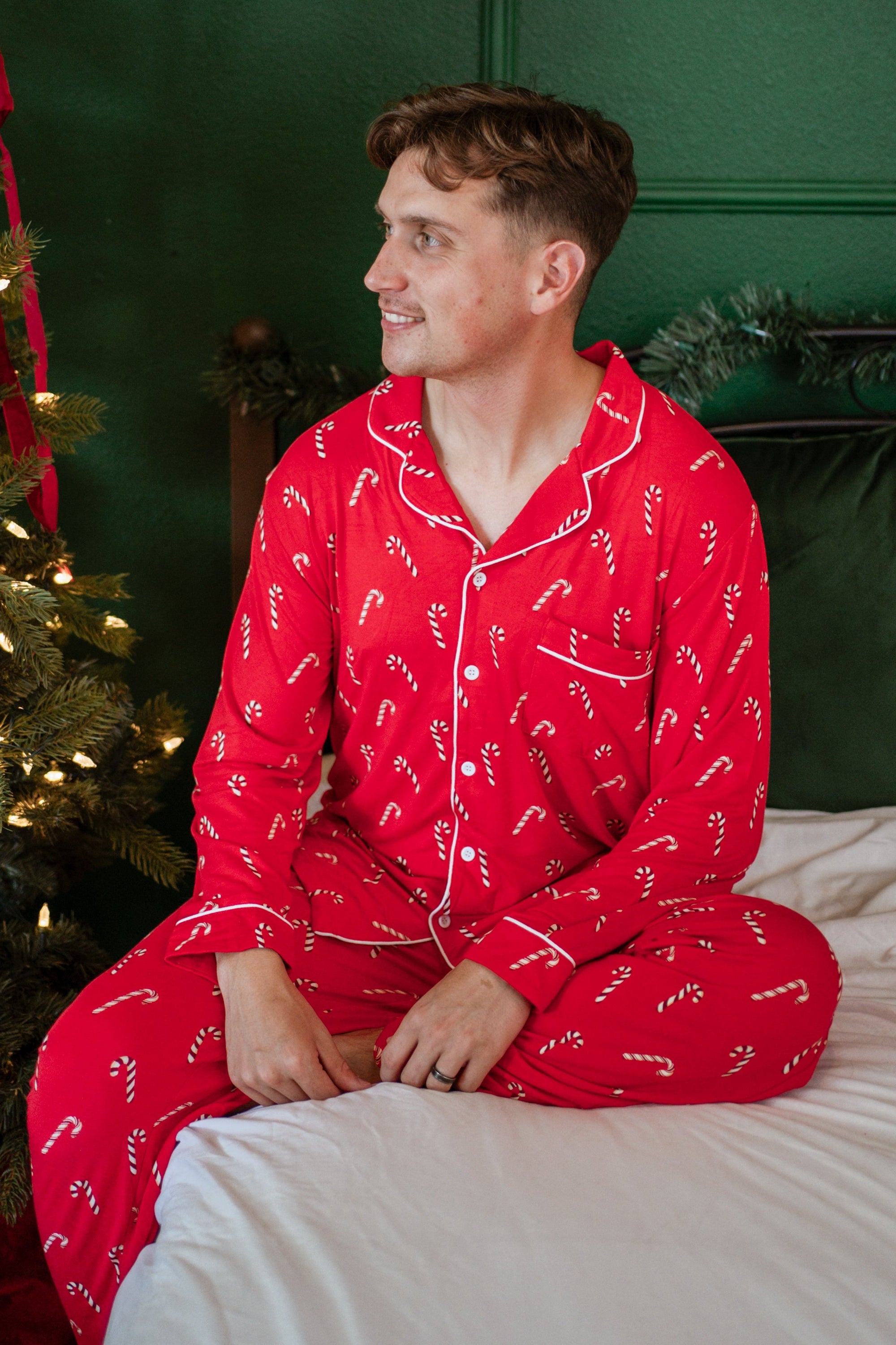 Male model sitting on a bed wearing the Men's Long Sleeve Pajama Set in Candy Cane with a decorated Christmas tree beside him