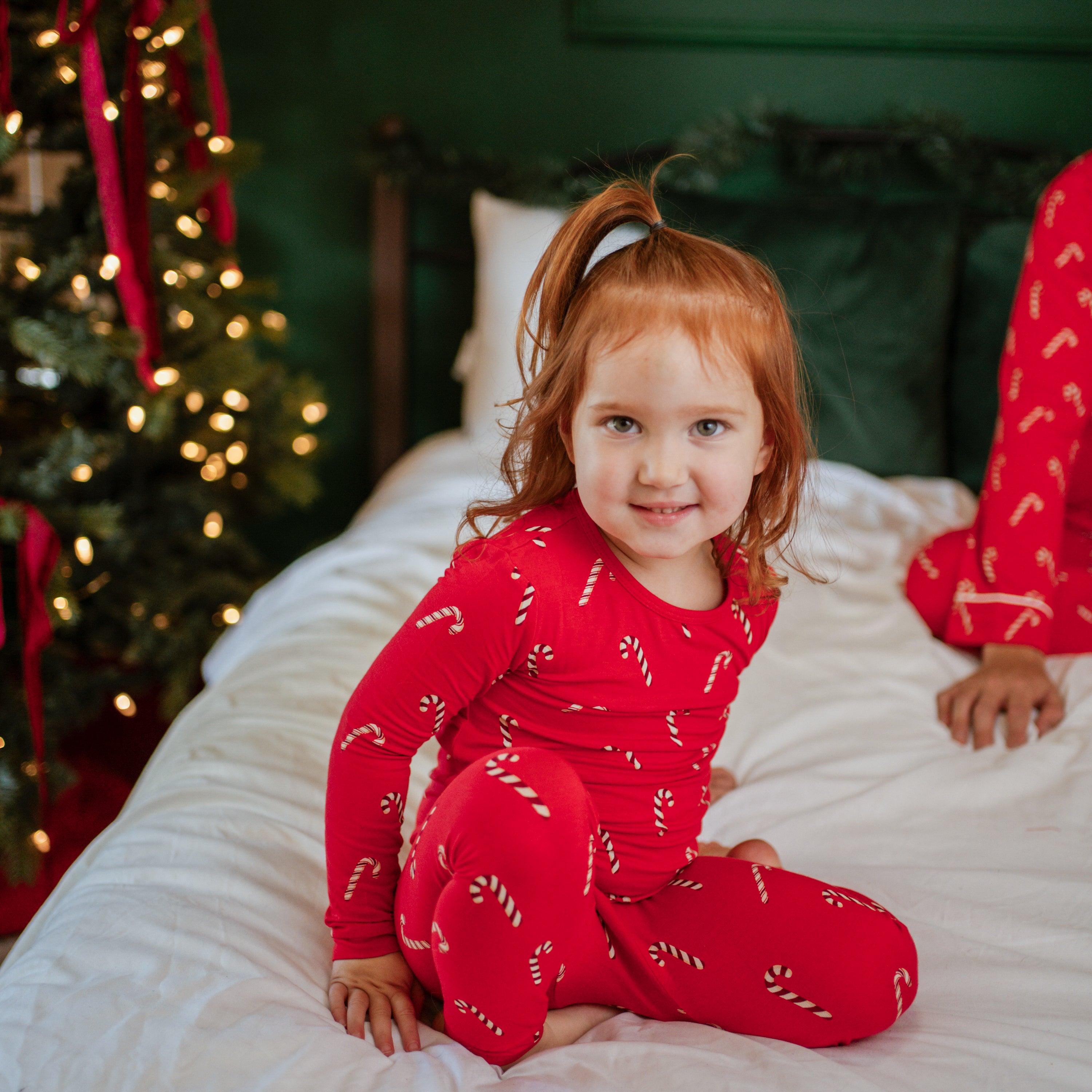 Smiling toddler sitting on a bed wearing the Long Sleeve Pajamas in Candy Cane
