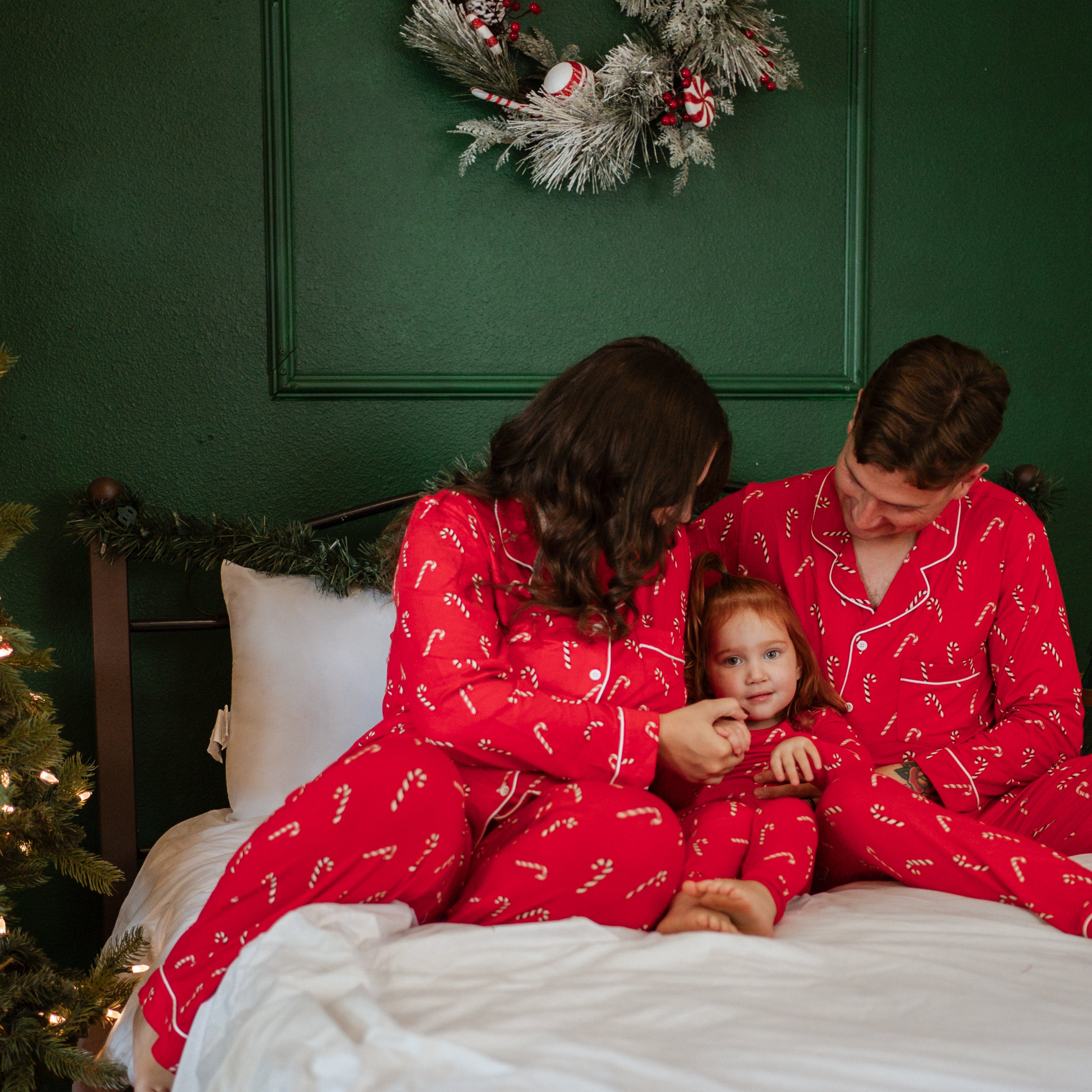 Family of three sitting on a bed all matching in the holiday print Candy Cane pajama sets
