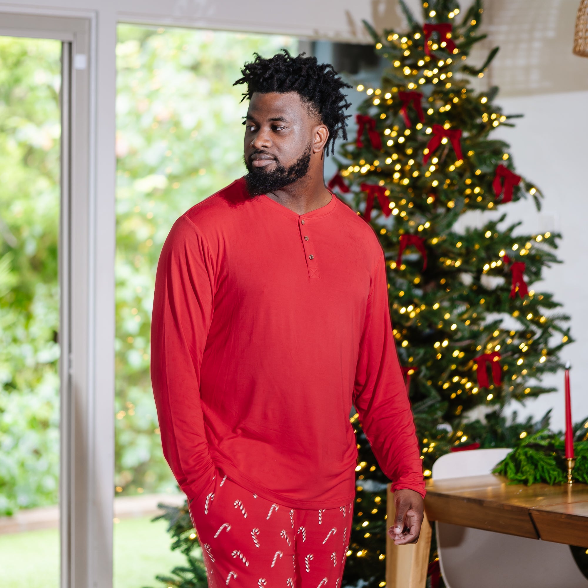 Male model standing in front of a Christmas tree wearing the Men's Long Sleeve Henley Top in Cardinal with Candy Cane Men's Lounge pants