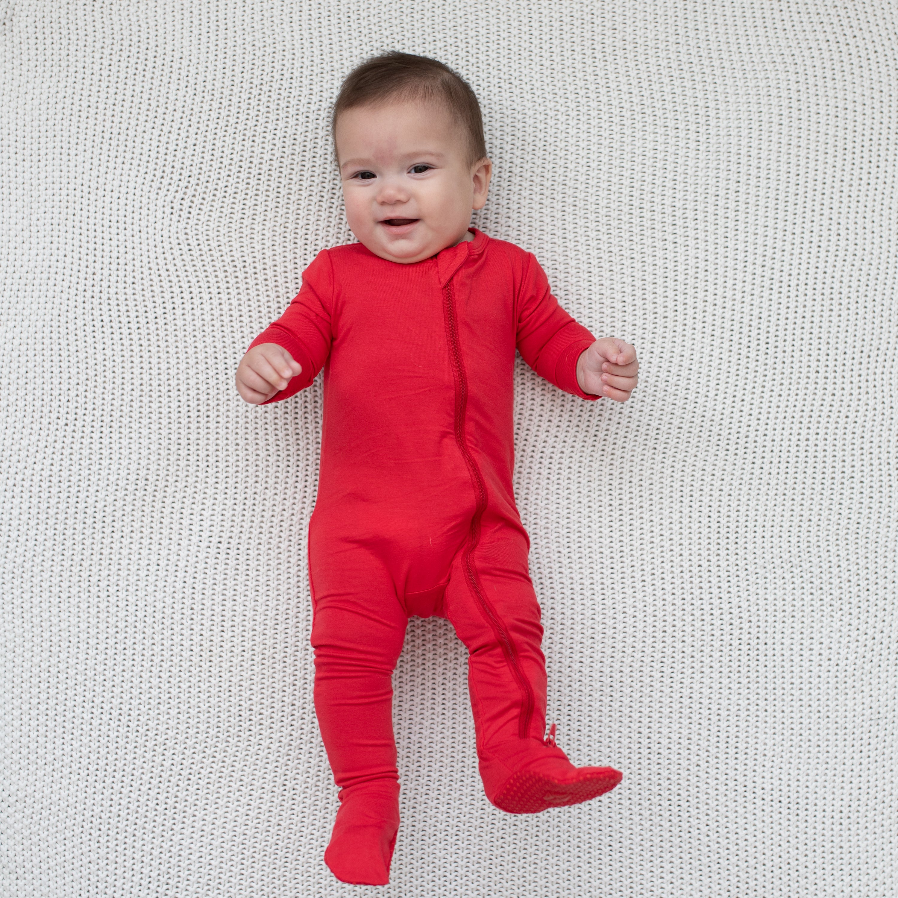 Baby modeling Zippered Footie in Cardinal laying on a knit blanket