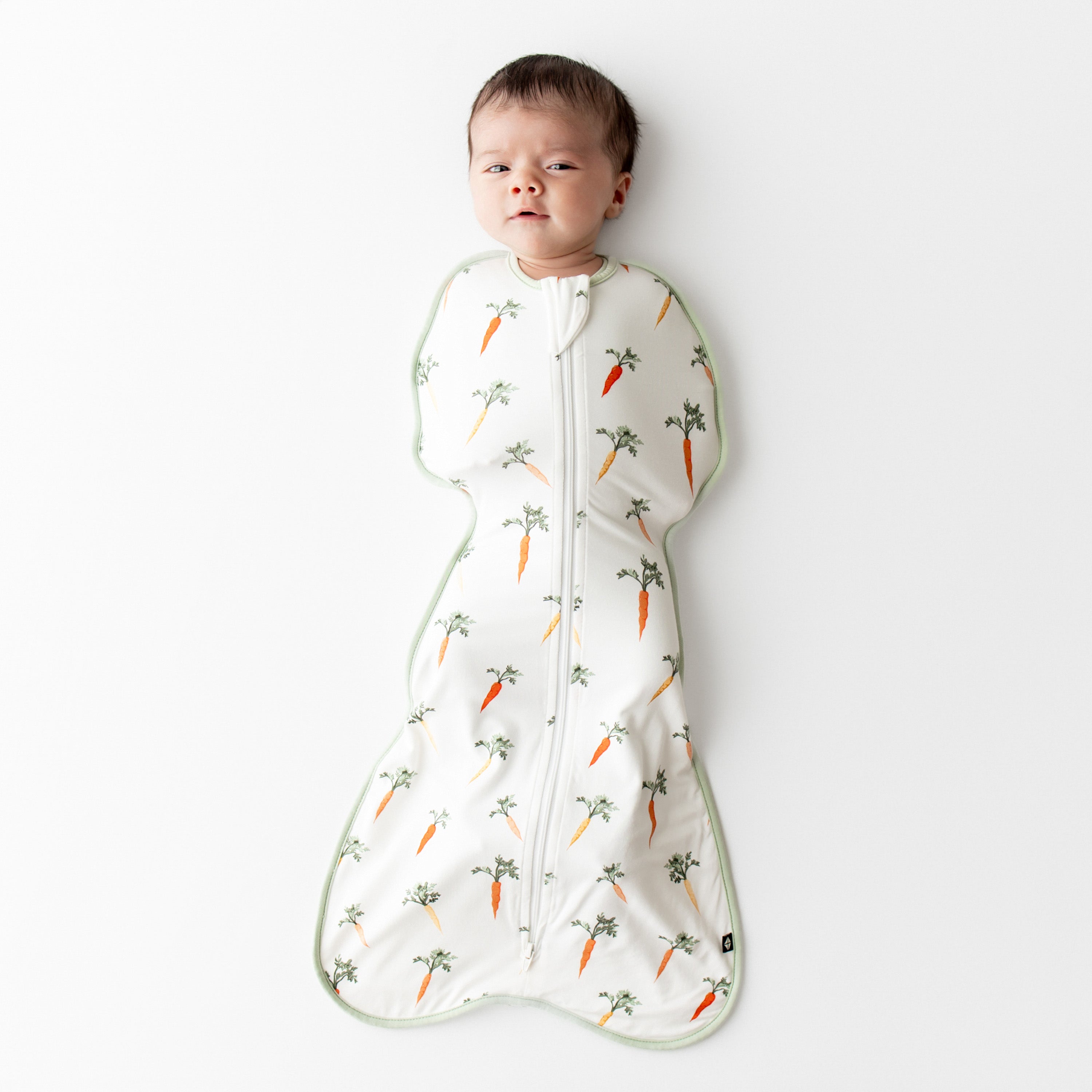Newborn laying on a white surface wearing the Swaddle Bag in Carrot 0.5