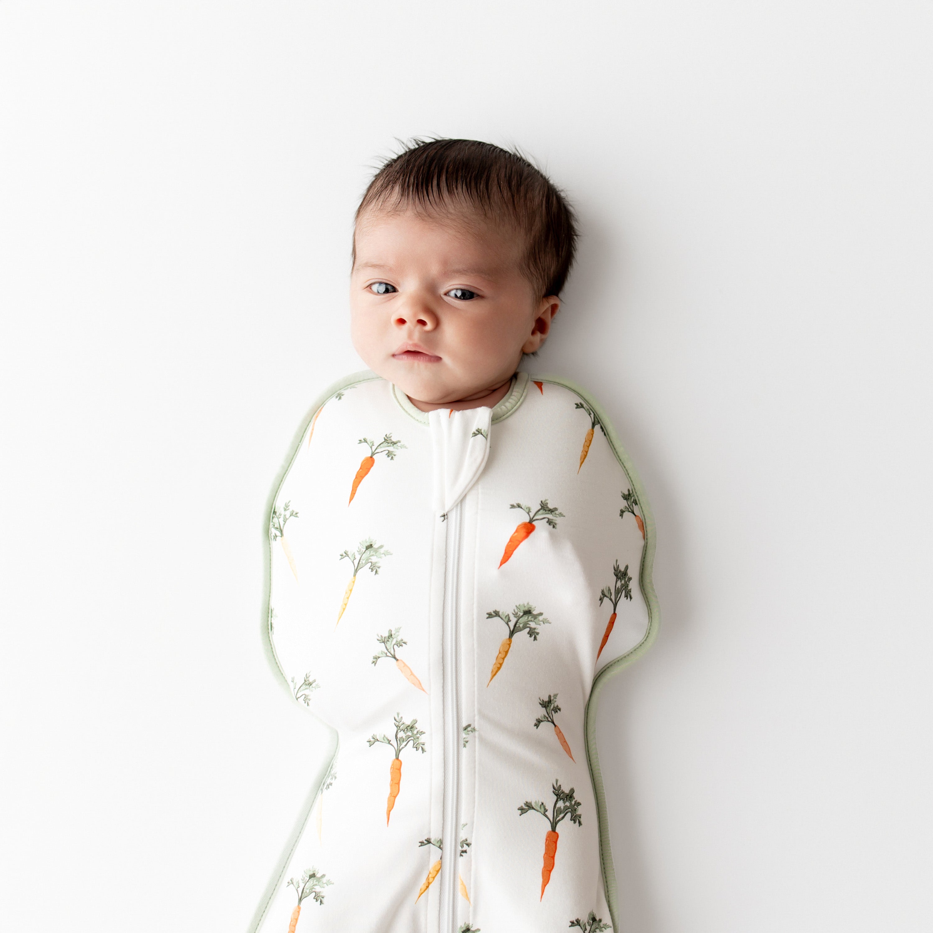 Close up of newborn wearing the Swaddle Bag in Carrot 0.5 laying on a white surface