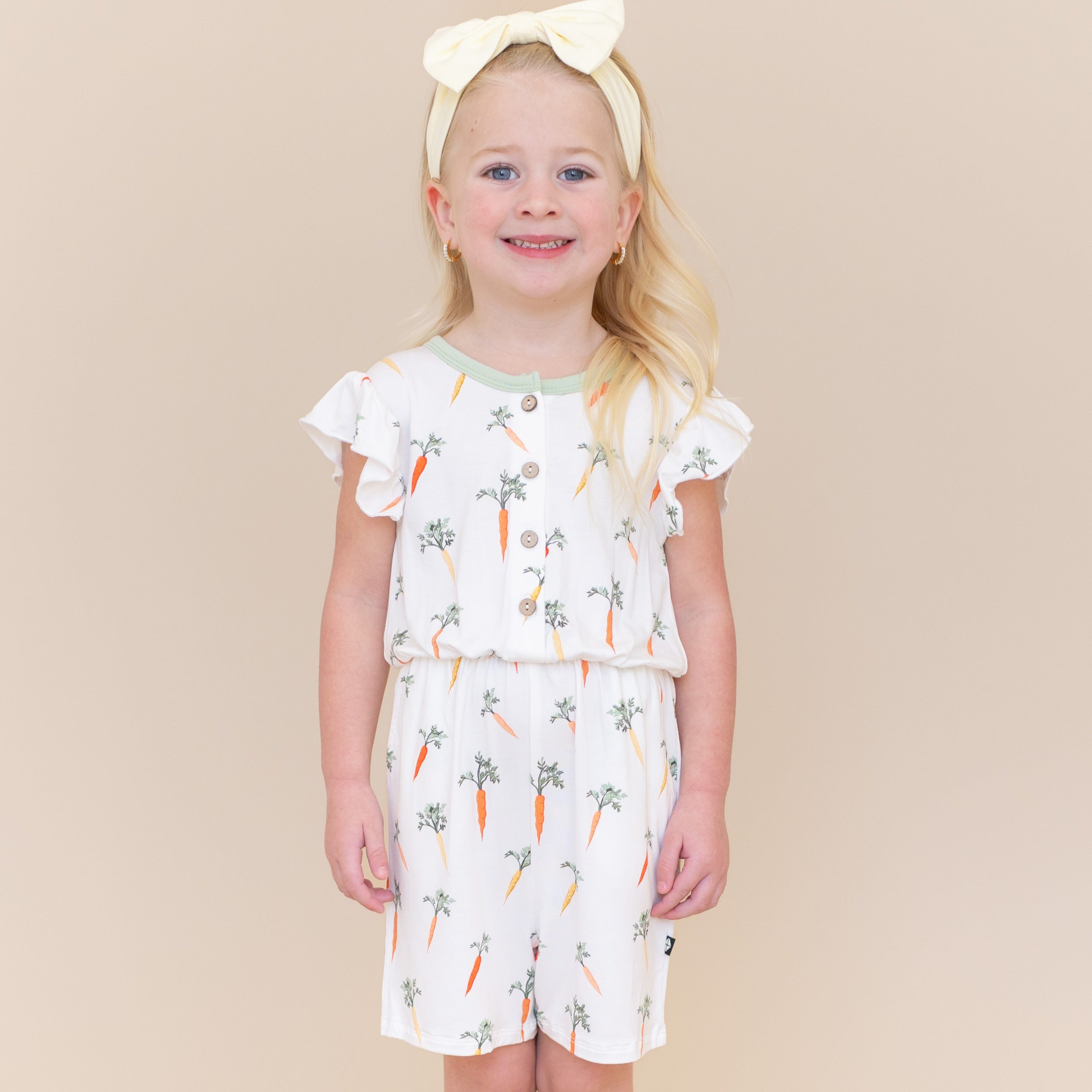 Young girl standing in front of a light taupe background wearing the Flutter Sleeve Romper in Carrot with a light yellow bow