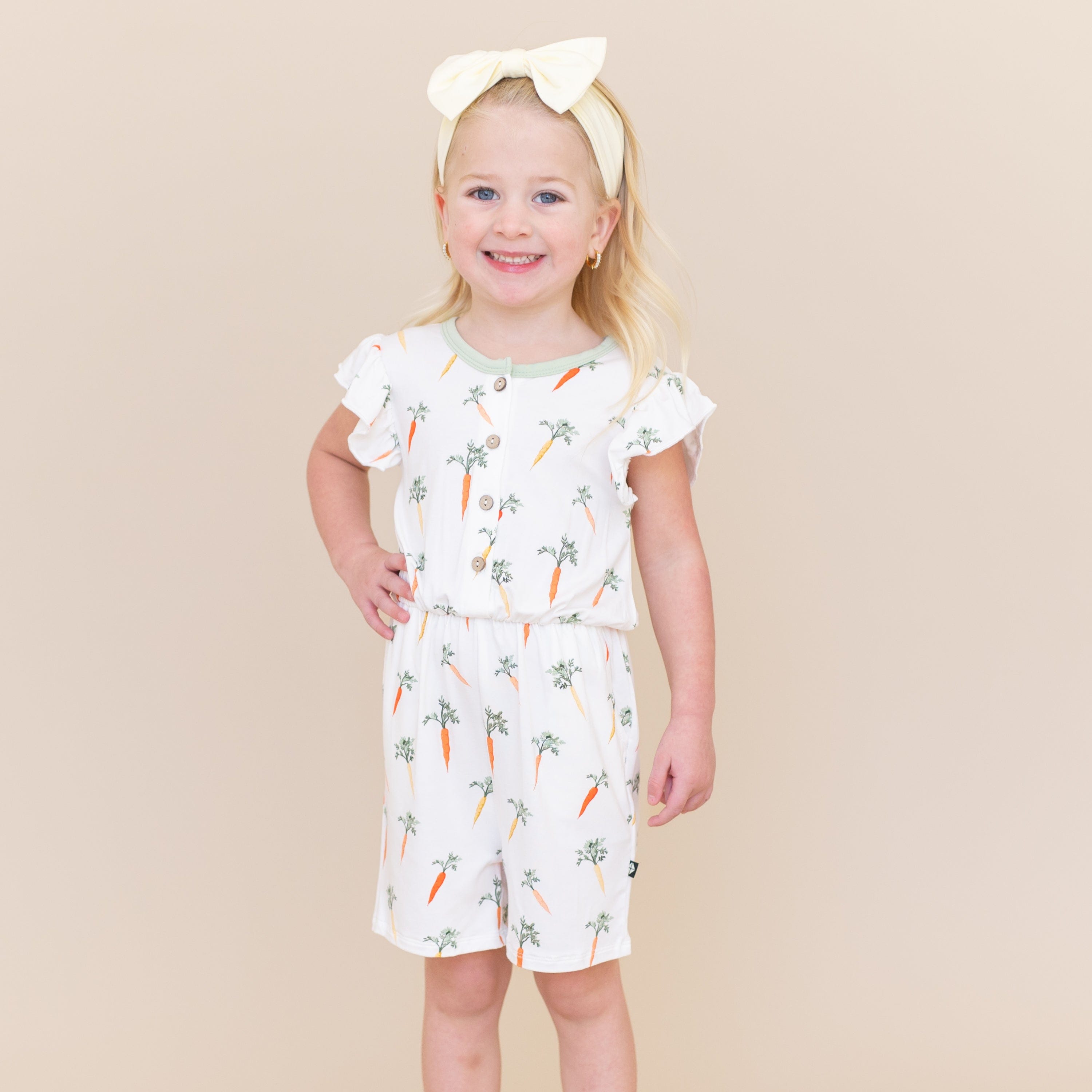 Young girl posing with one hand on her hip wearing the Flutter Sleeve Romper in Carrot with a light yellow bow