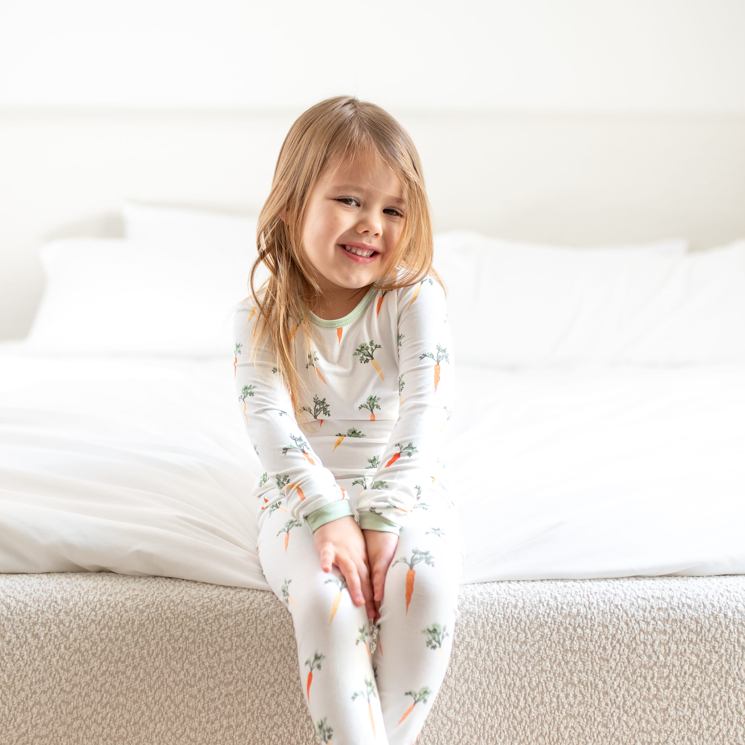 Young girl sitting at the end of a bed with her hands on the top of her knees wearing the Long Sleeve Pajamas in Carrot