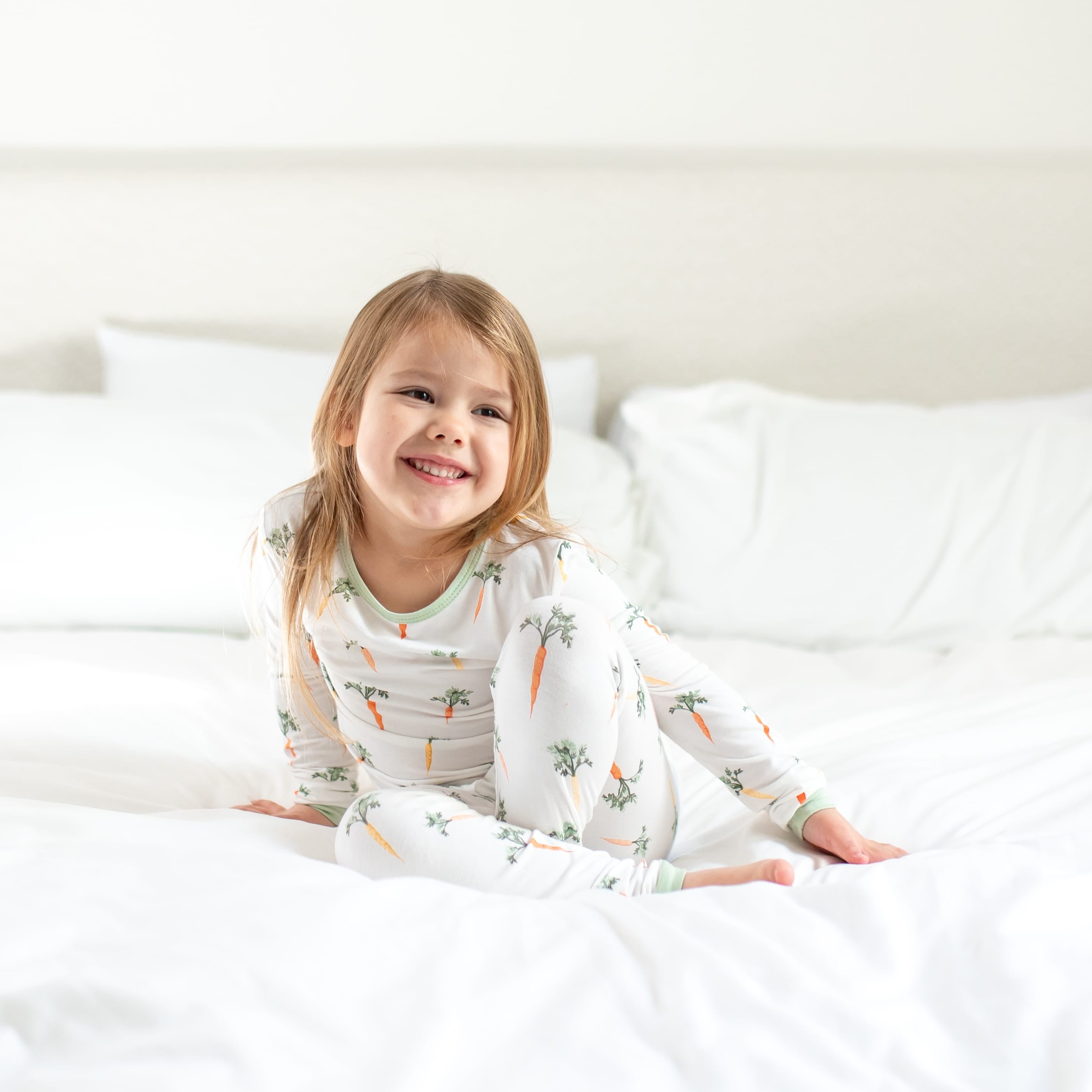 Young smiling girl sitting in a bed wearing the Long Sleeve Pajamas in Carrot