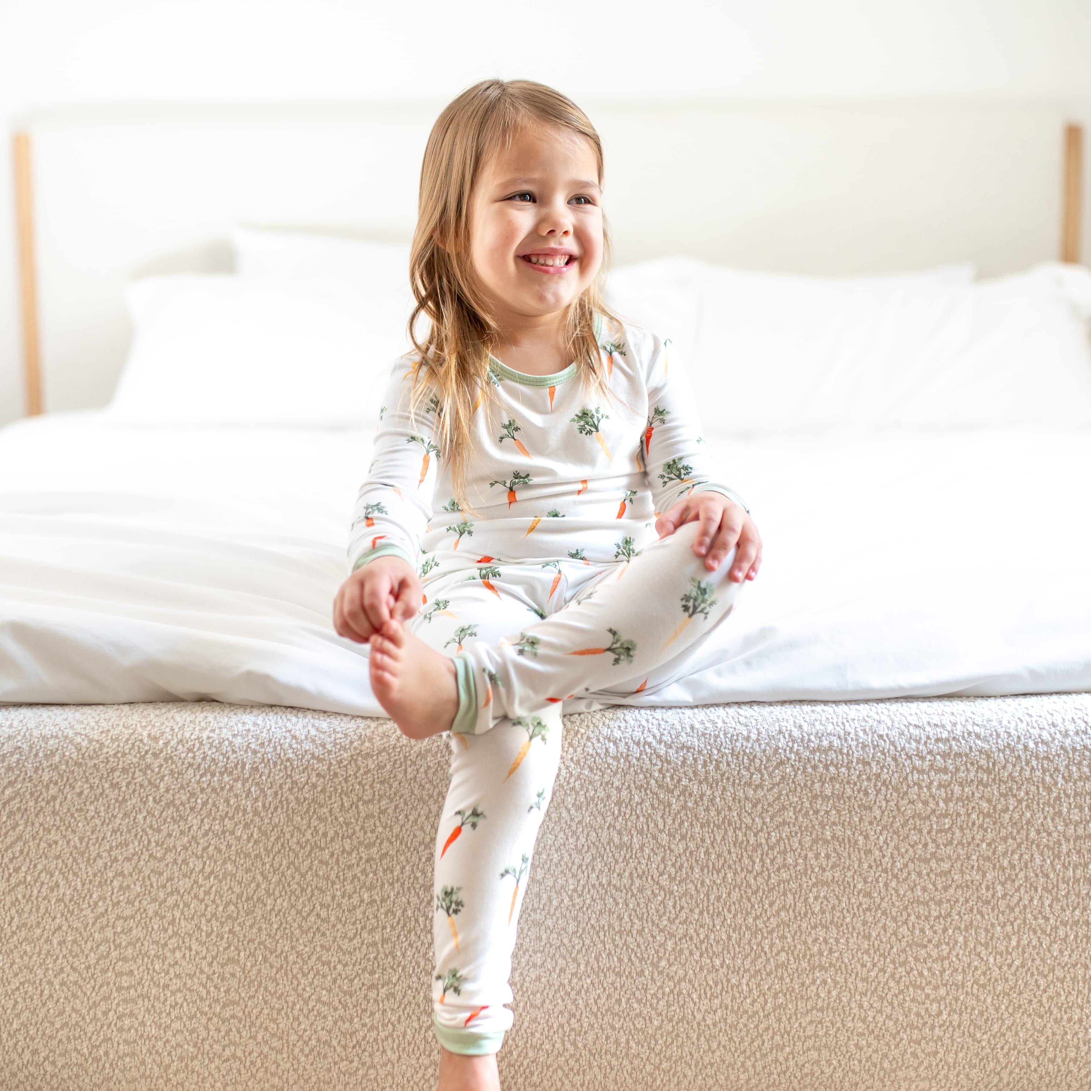 Young girl sitting at the edge of the bed wearing the Long Sleeve Pajamas in Carrot
