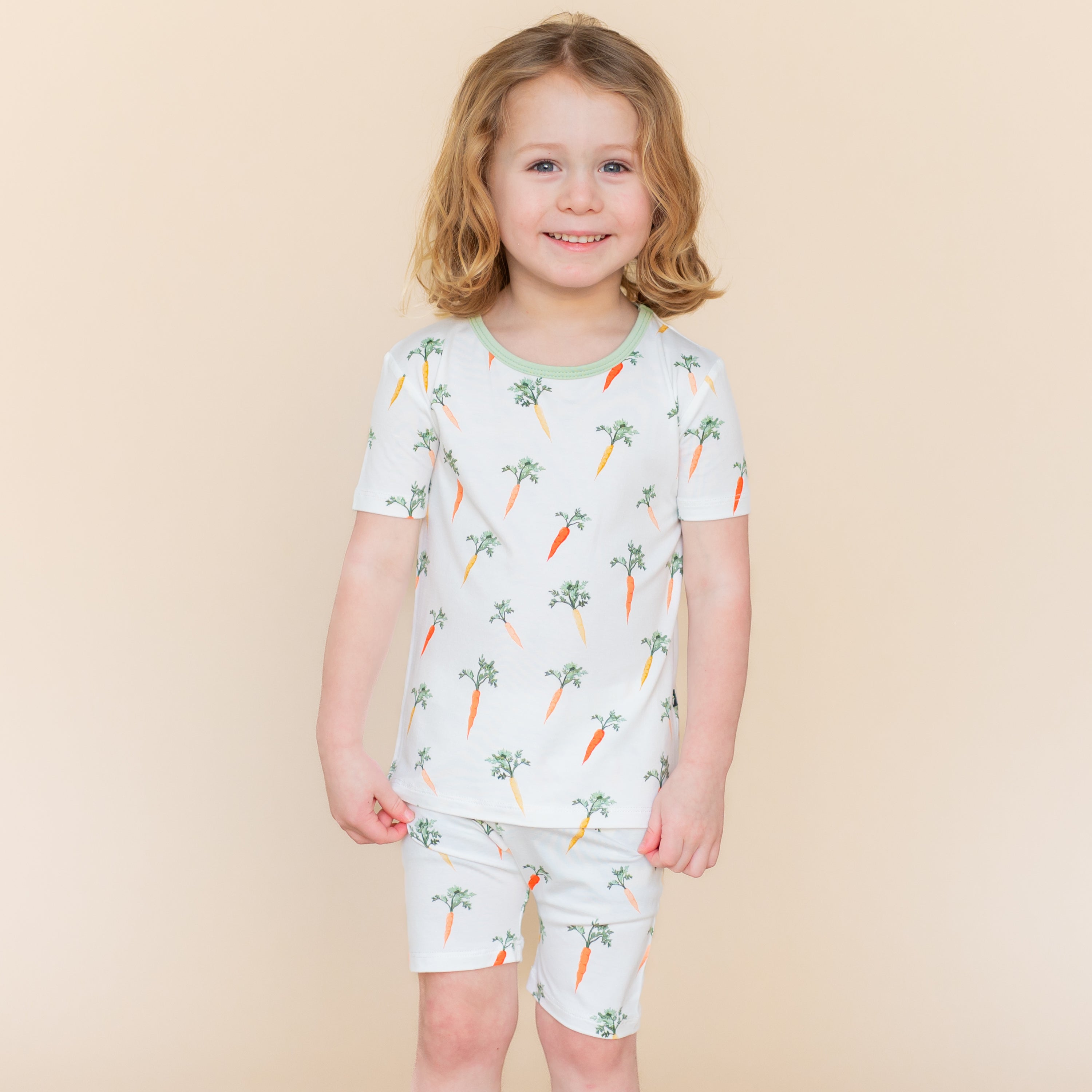 Young smiling child modeling the Short Sleeve Pajamas in Carrot standing in front of a light taupe background