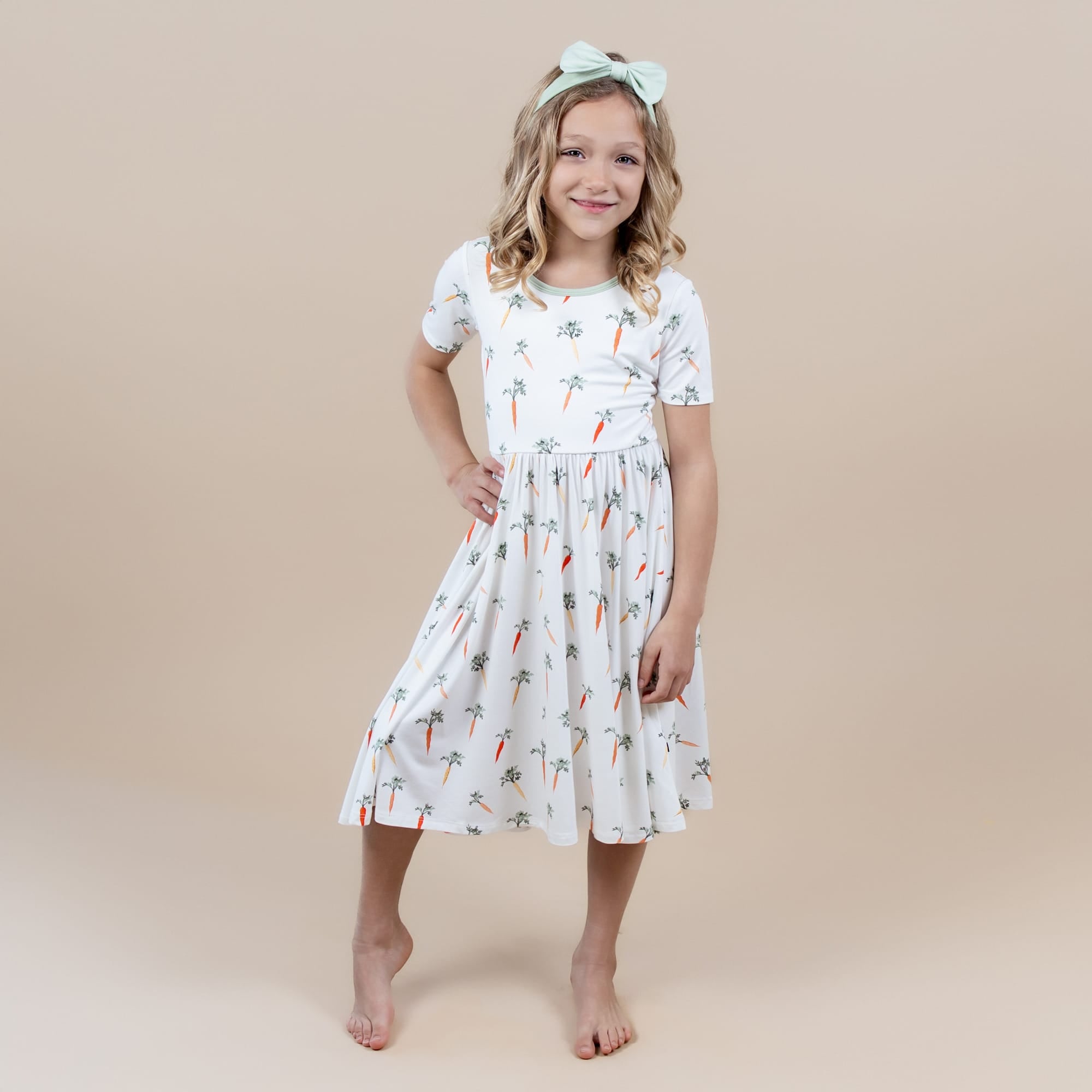 Young girl posing wearing the Twirl Dress in Carrot paired with a light green bow