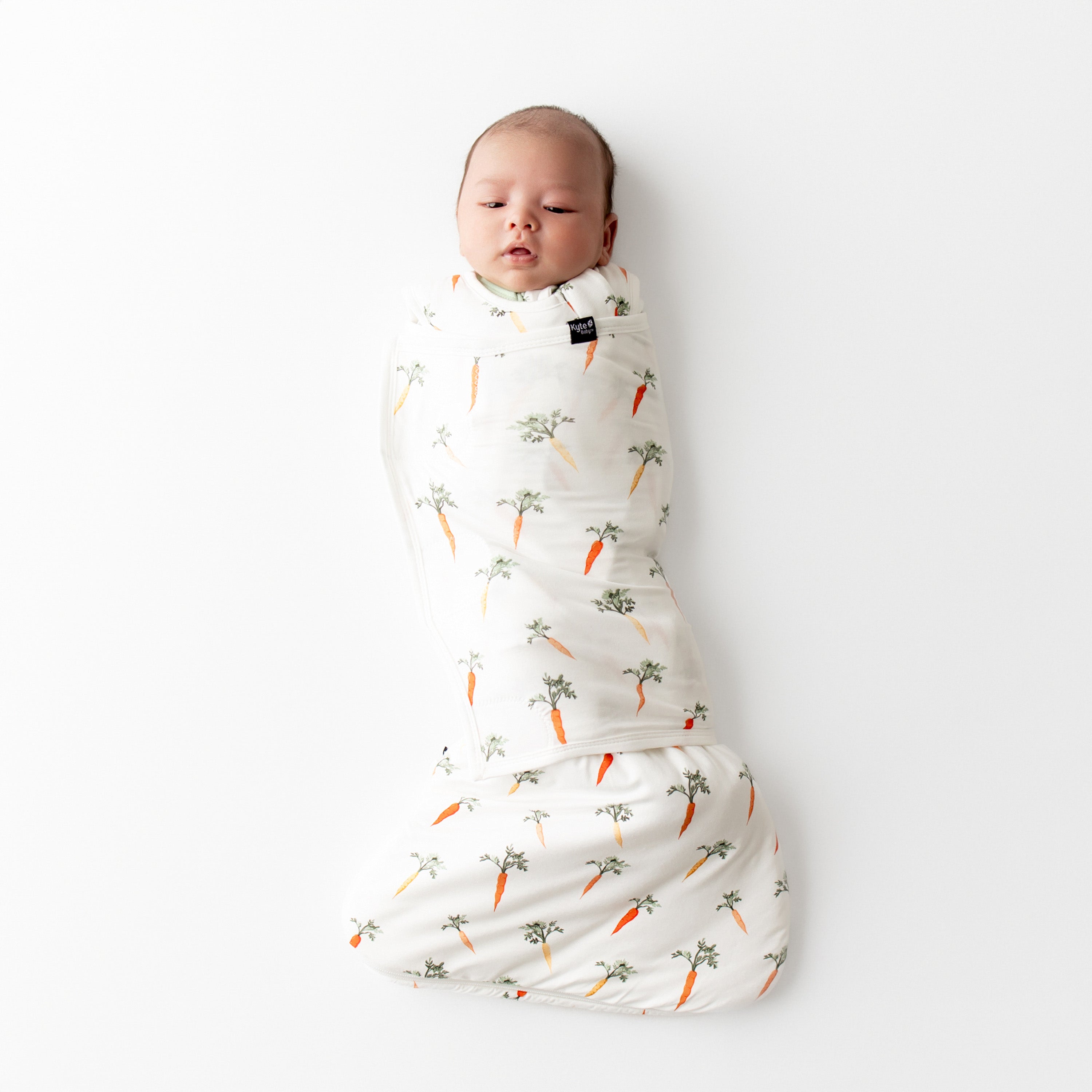 Newborn baby swaddled in the Sleep Bag Swaddler in Carrot 1.0 laying on a white surface