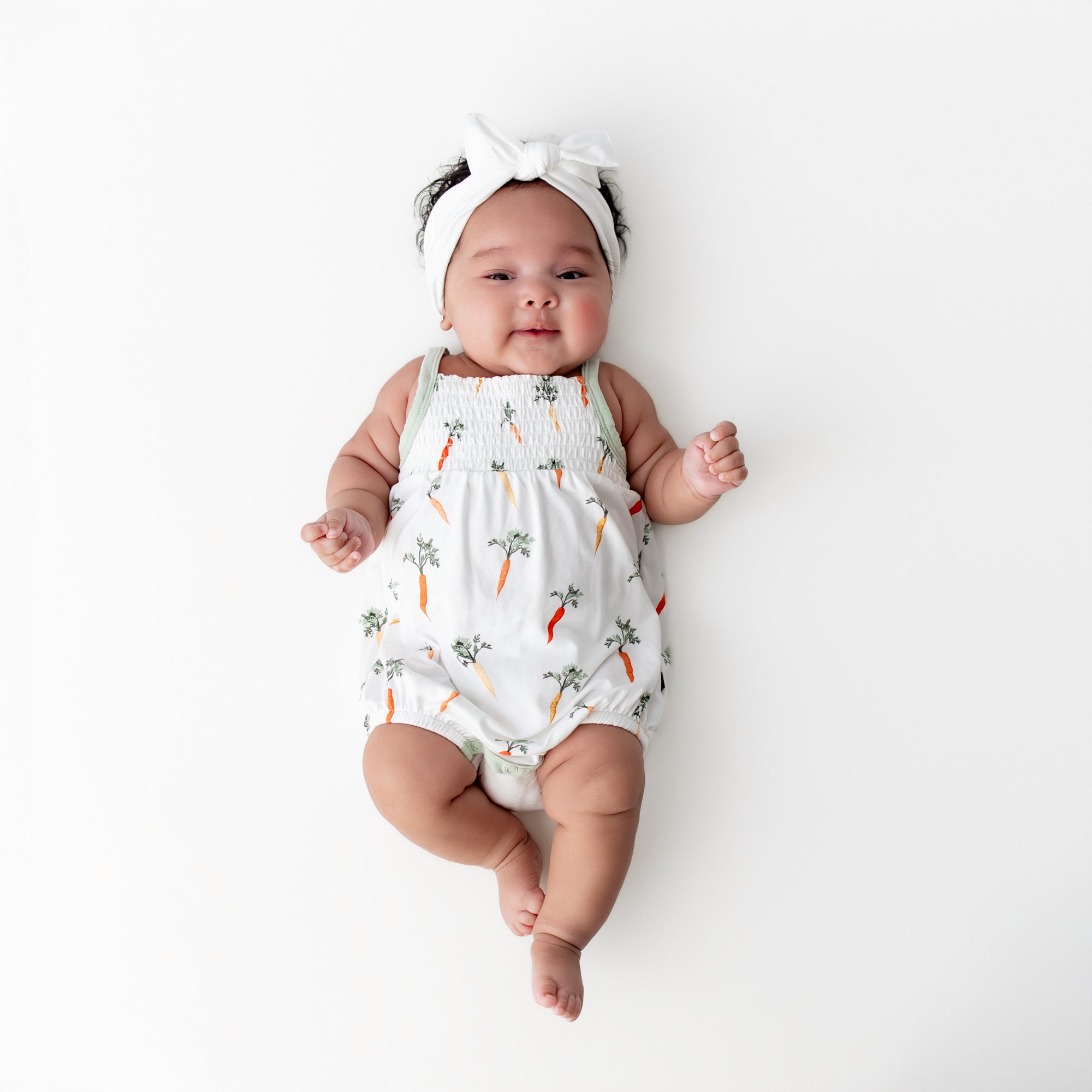 Infant laying on a white surface wearing the Smocked Bubble Romper in Carrot with a white bow