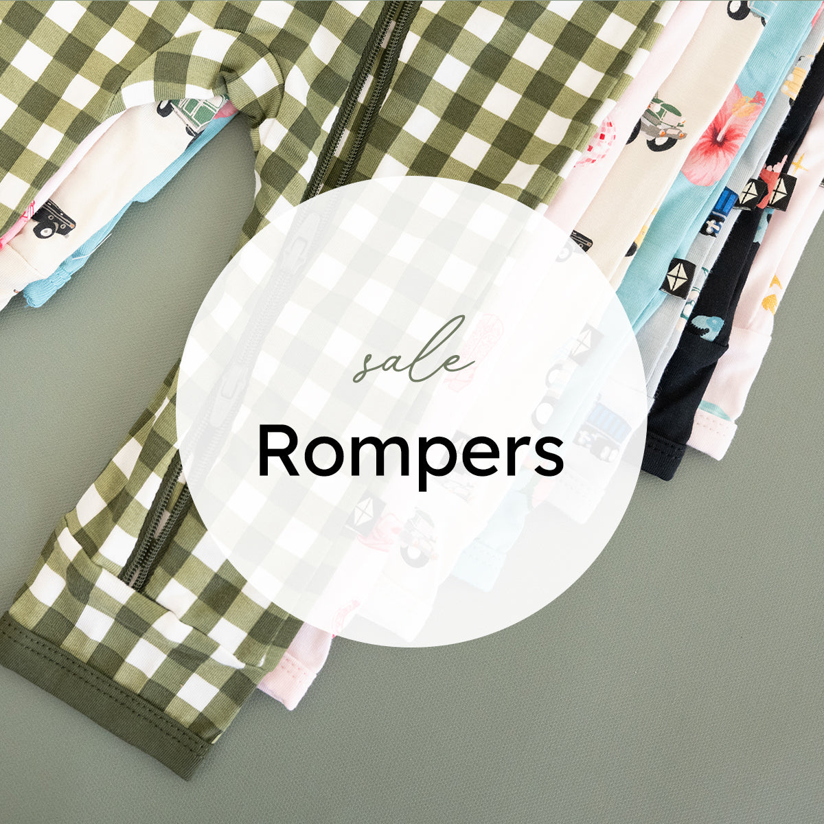 A promotional flat-lay showing several children's zip-front rompers stacked diagonally on a gray surface. A large white oval overlay in the center reads 'sale Rompers' in black text.