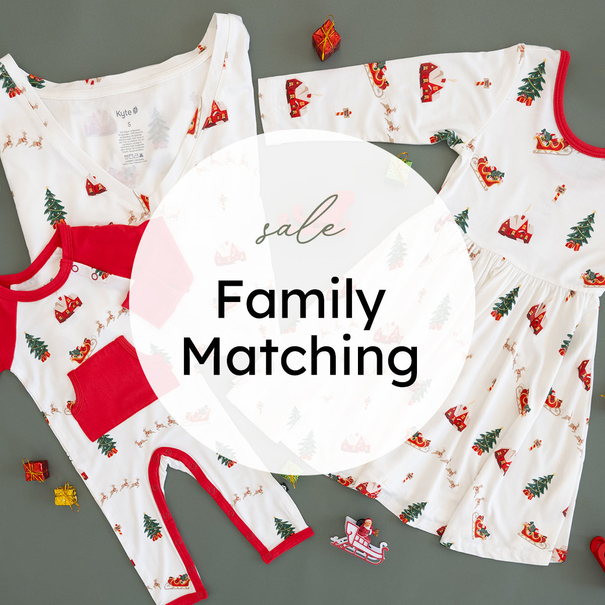 A flat lay of a Christmas-themed family matching pajama set on a gray background, featuring a white long-sleeve top and matching pants with red trim and a festive print of Santa Claus, Christmas trees, and reindeer. A matching white and red long-sleeve dress is also shown.