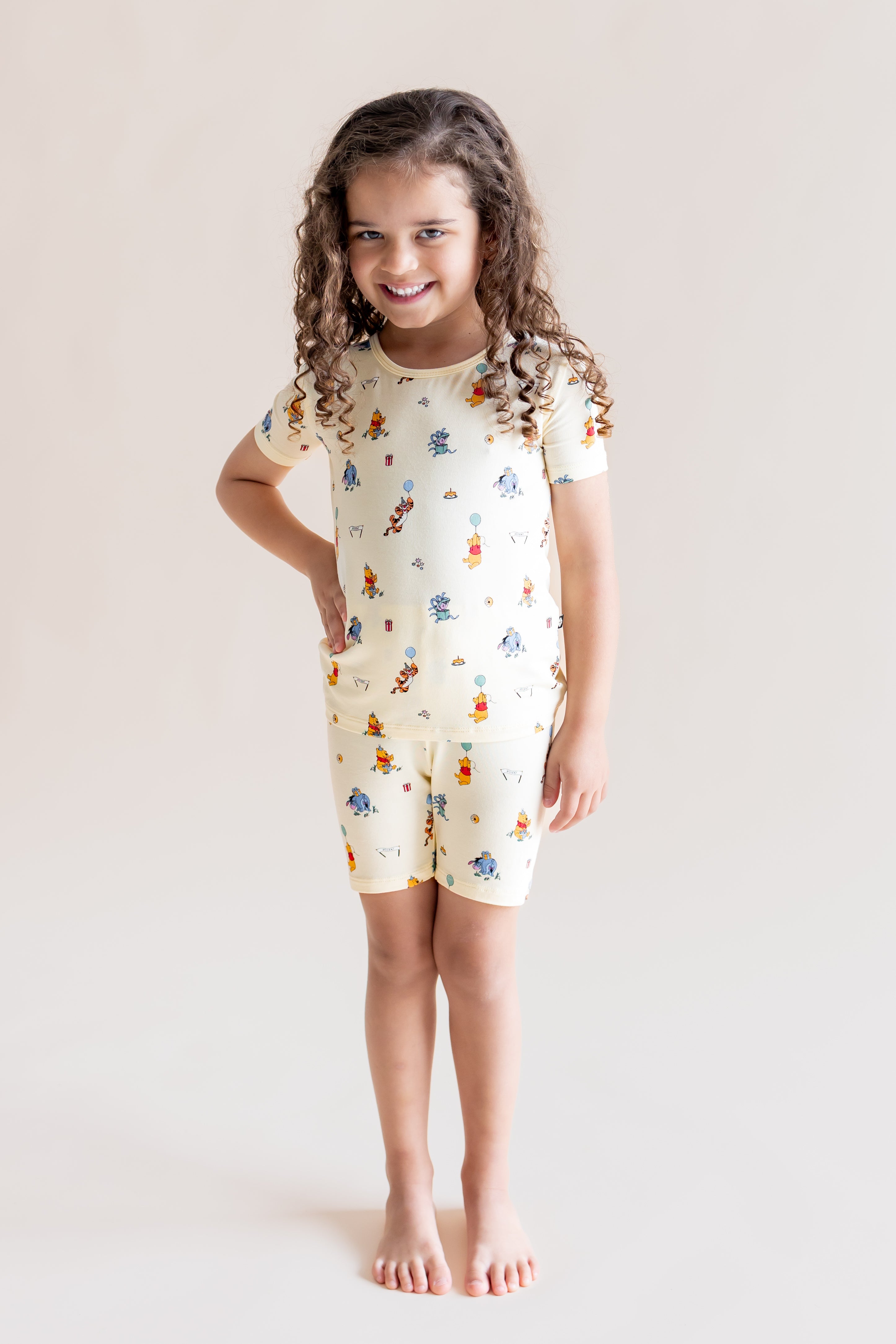Girl wearing Short Sleeve Pajamas in Winnie the Pooh Celebration print posing on a cream background