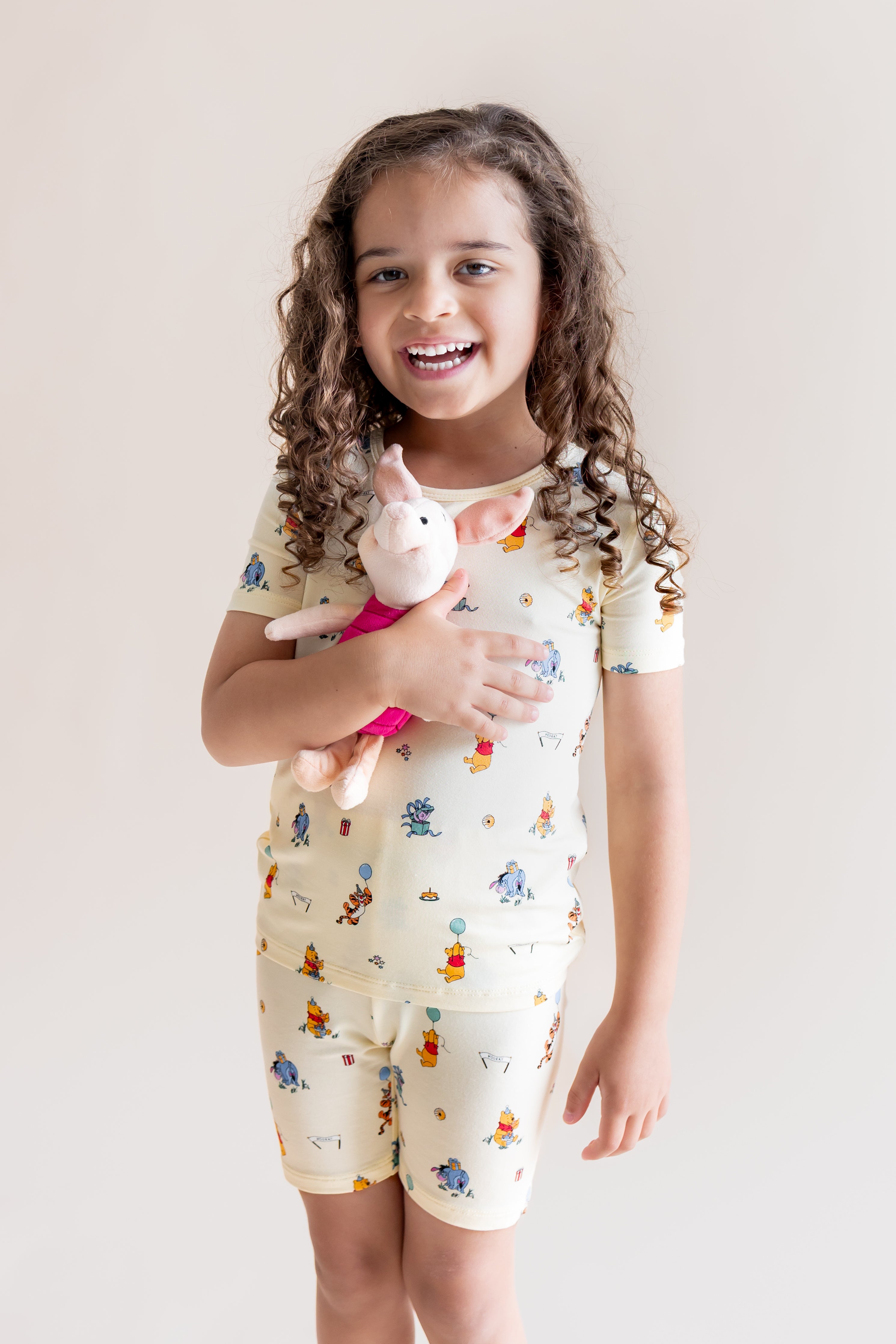 Girl holding a stuff toy wearing Short Sleeve Pajamas in Winnie the Pooh Celebration print on a cream background